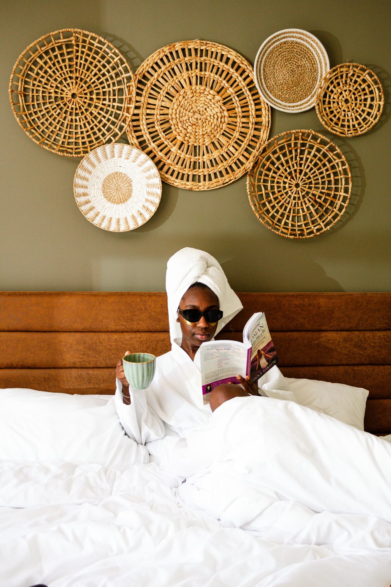 Person in white towel and sunglasses reading a book and holding a cup in bed, with woven baskets on the wall behind.