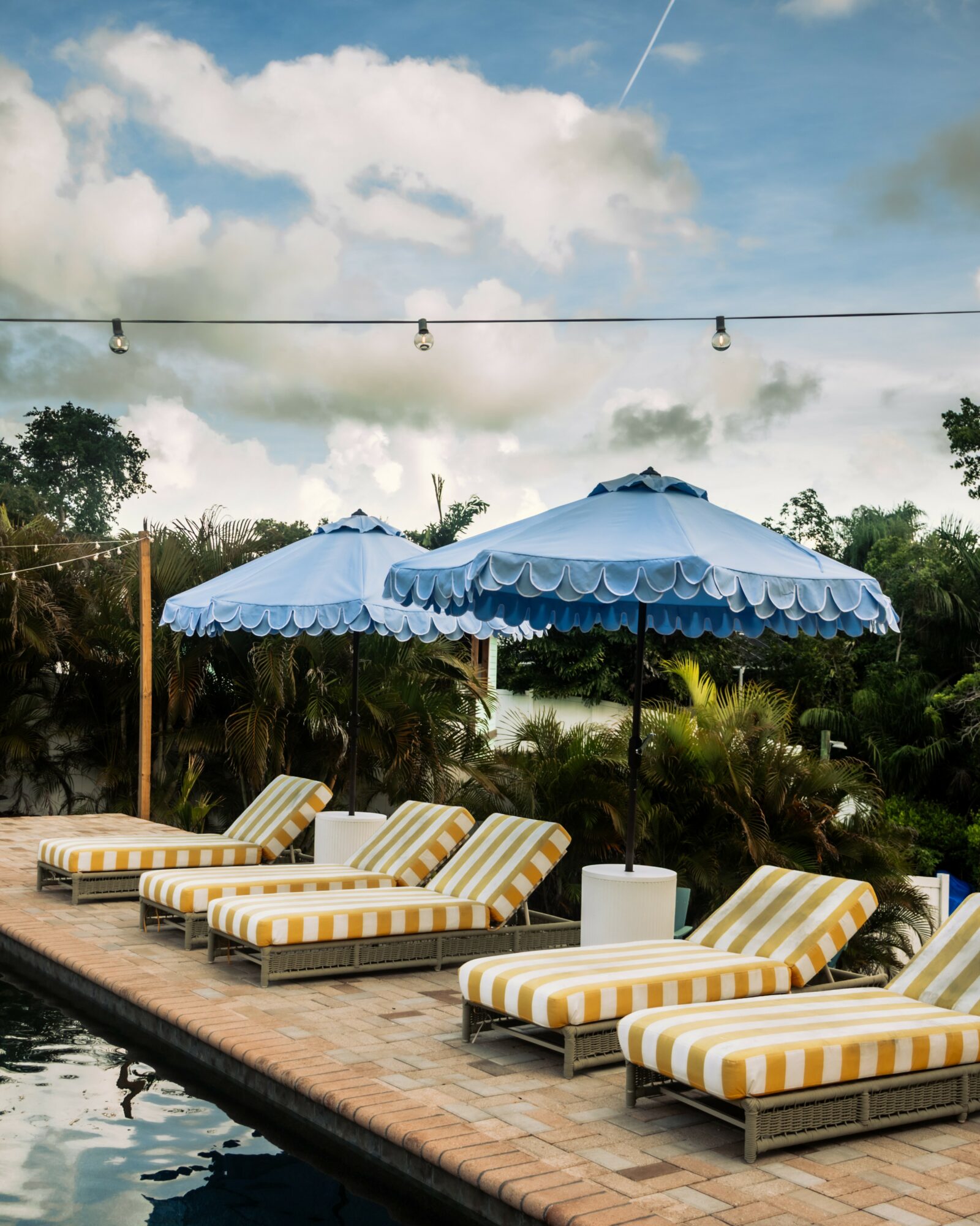 Poolside area with striped lounge chairs, blue umbrellas, and string lights under a partly cloudy sky.