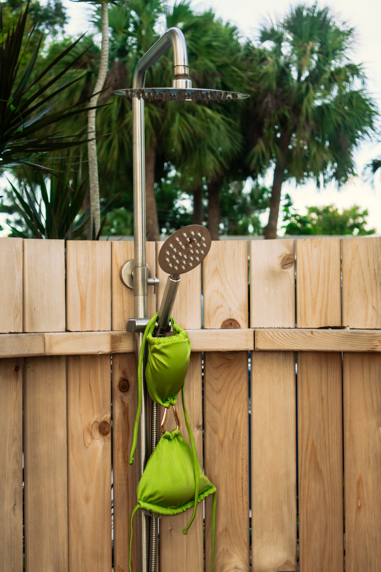 Outdoor shower with a green bag hanging, wooden fence, and palm trees in background.
