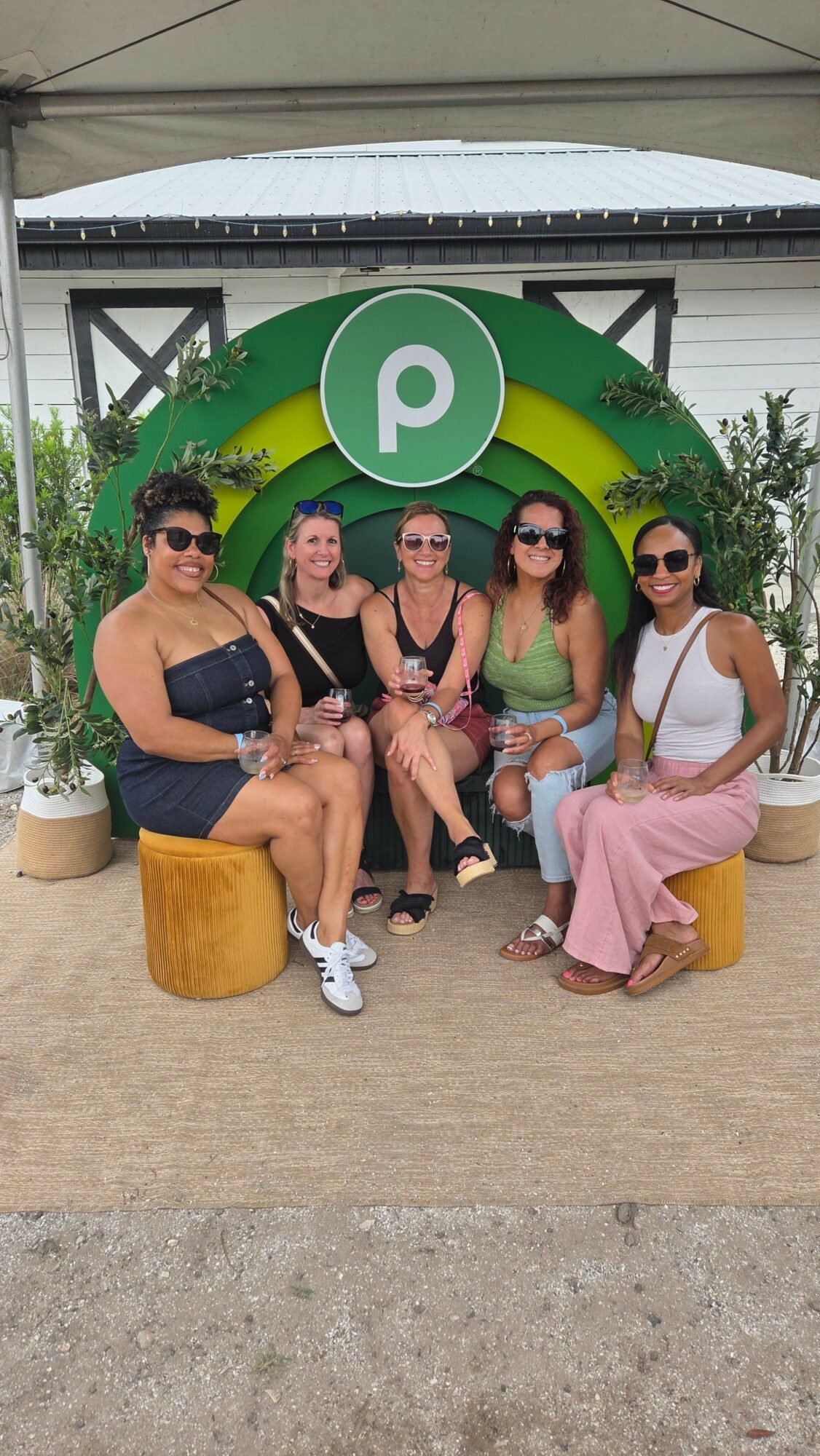 Group of six women sitting and standing outdoors in front of a green and white sign, some holding drinks, smiling.