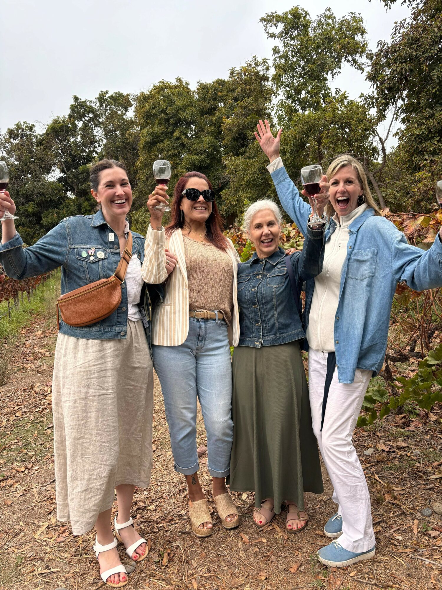 Four women standing outdoors, smiling, holding glasses, with trees in the background.