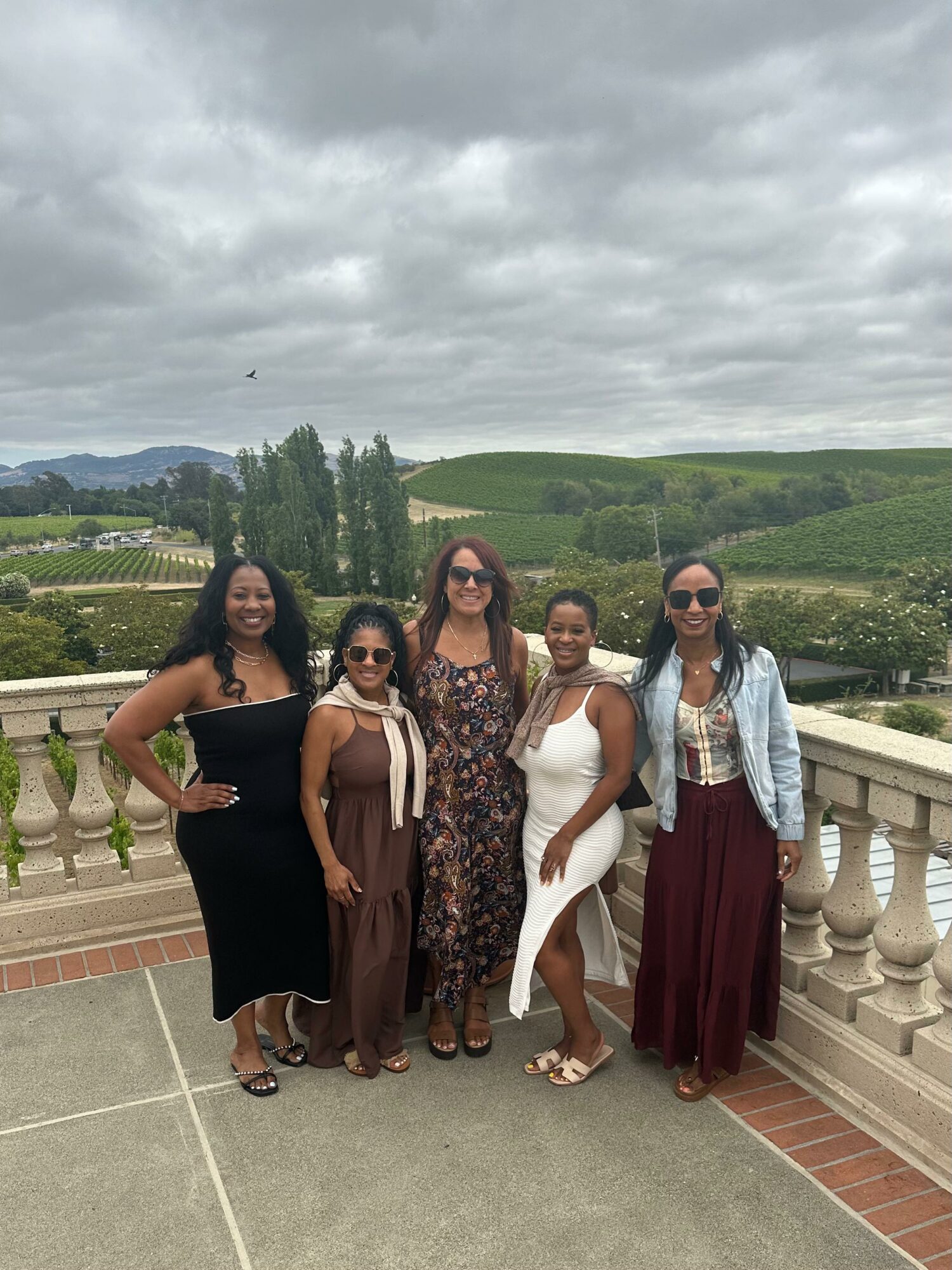 Five women standing on a balcony with a scenic landscape of rolling hills and cloudy sky in the background.