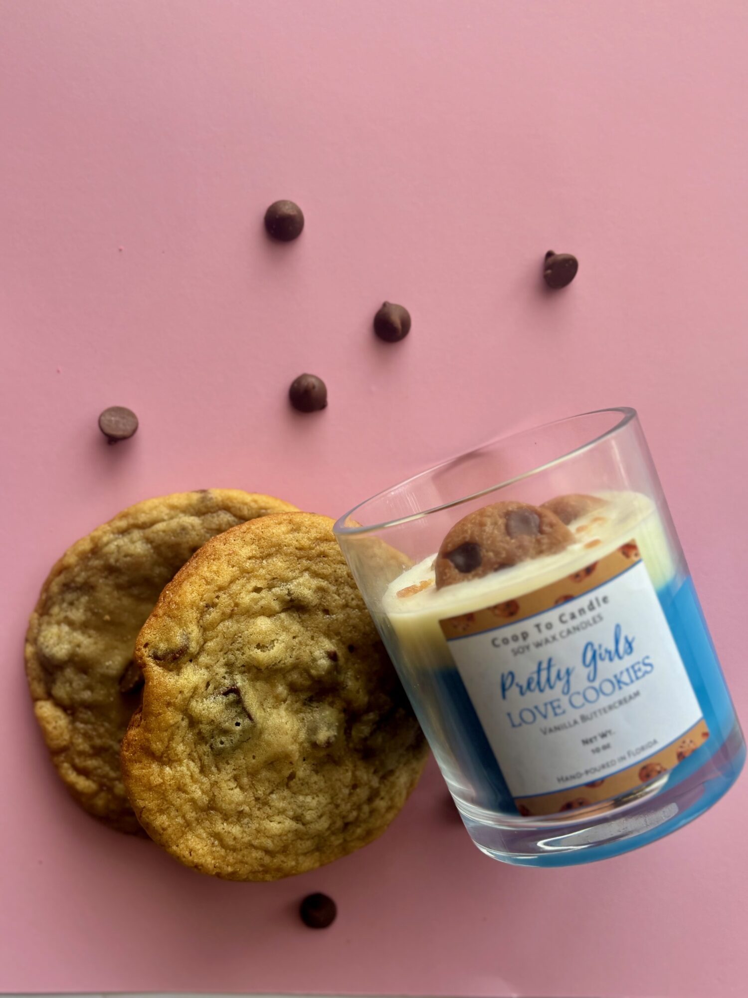 Two cookies and a glass of milk with cookies on a pink background, scattered chocolate chips, and a cookie in the glass.
