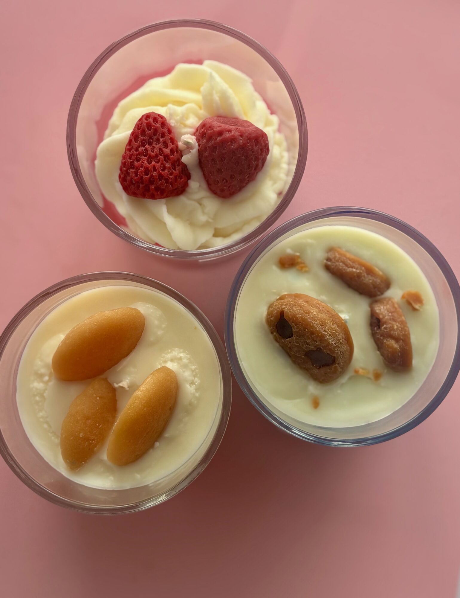 Three glasses of dessert with strawberries, dried fruits, and cream on a pink surface.