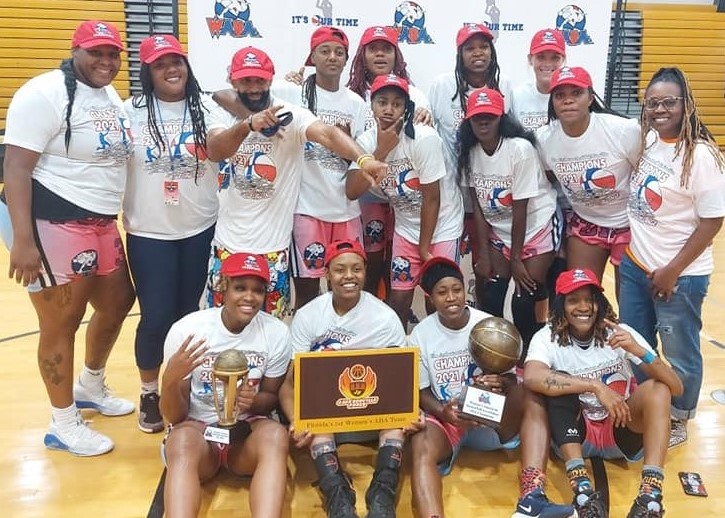 Group of young girls in sports uniforms and caps, holding trophies and medals, celebrating indoors with a basketball court background.