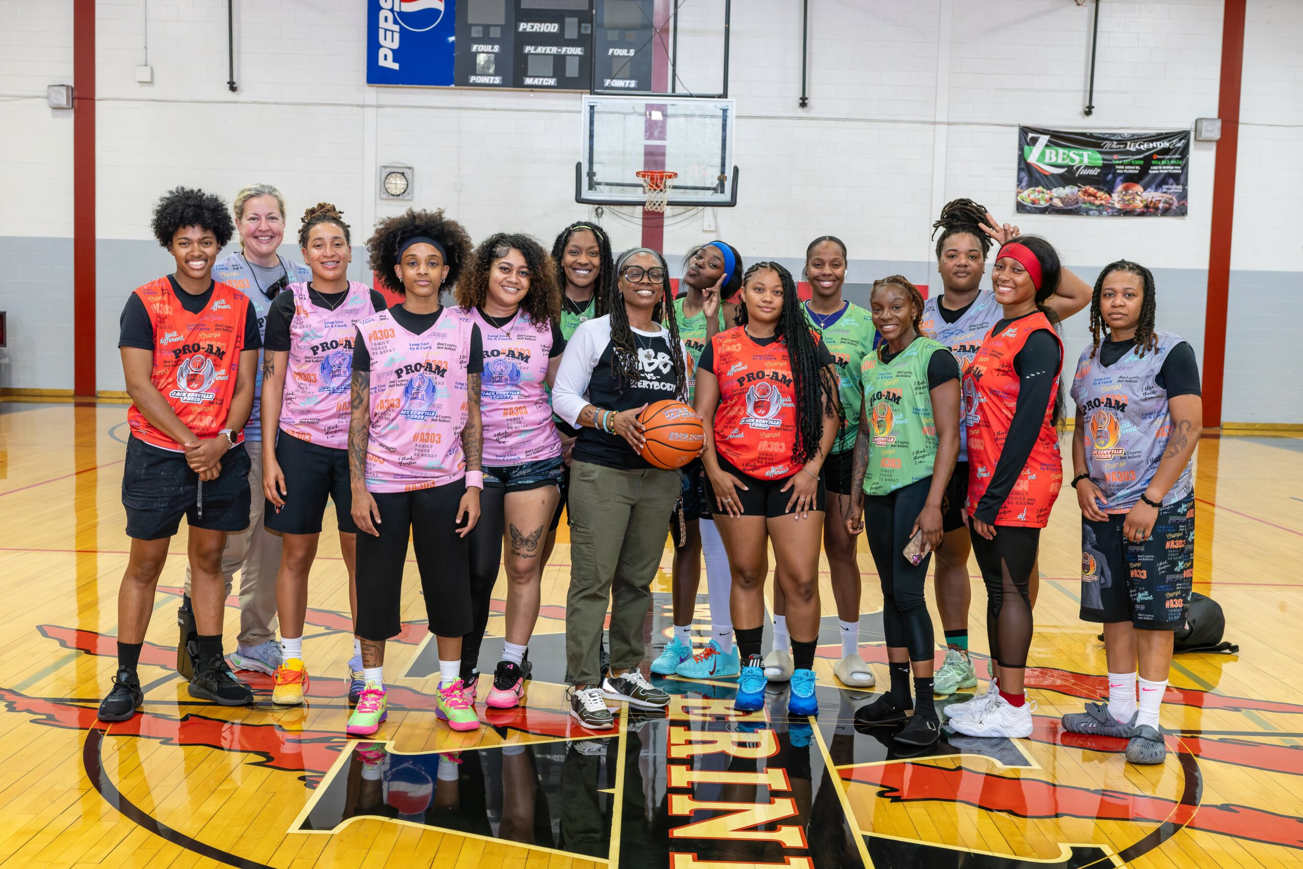 Group of young people in a gymnasium, some holding a basketball, standing in front of a basketball hoop, smiling.