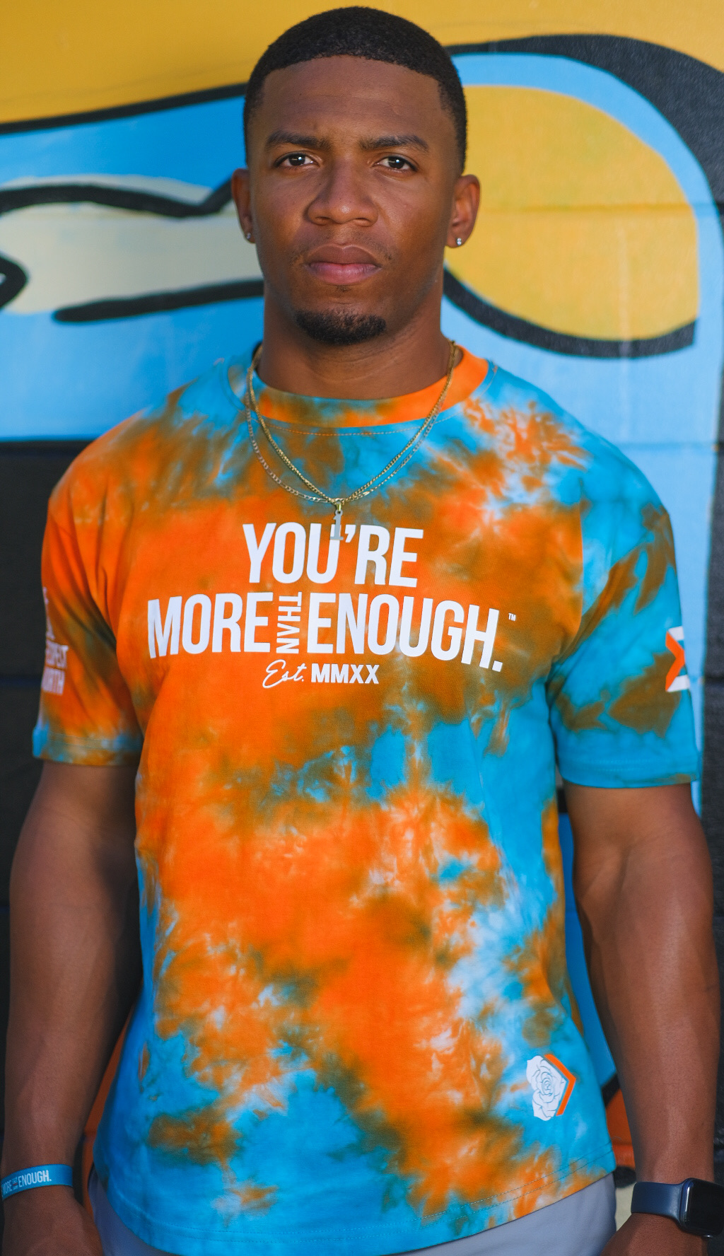 Young man with short hair and a serious expression, wearing a colorful tie-dye shirt with text, standing in front of a graffiti wall.