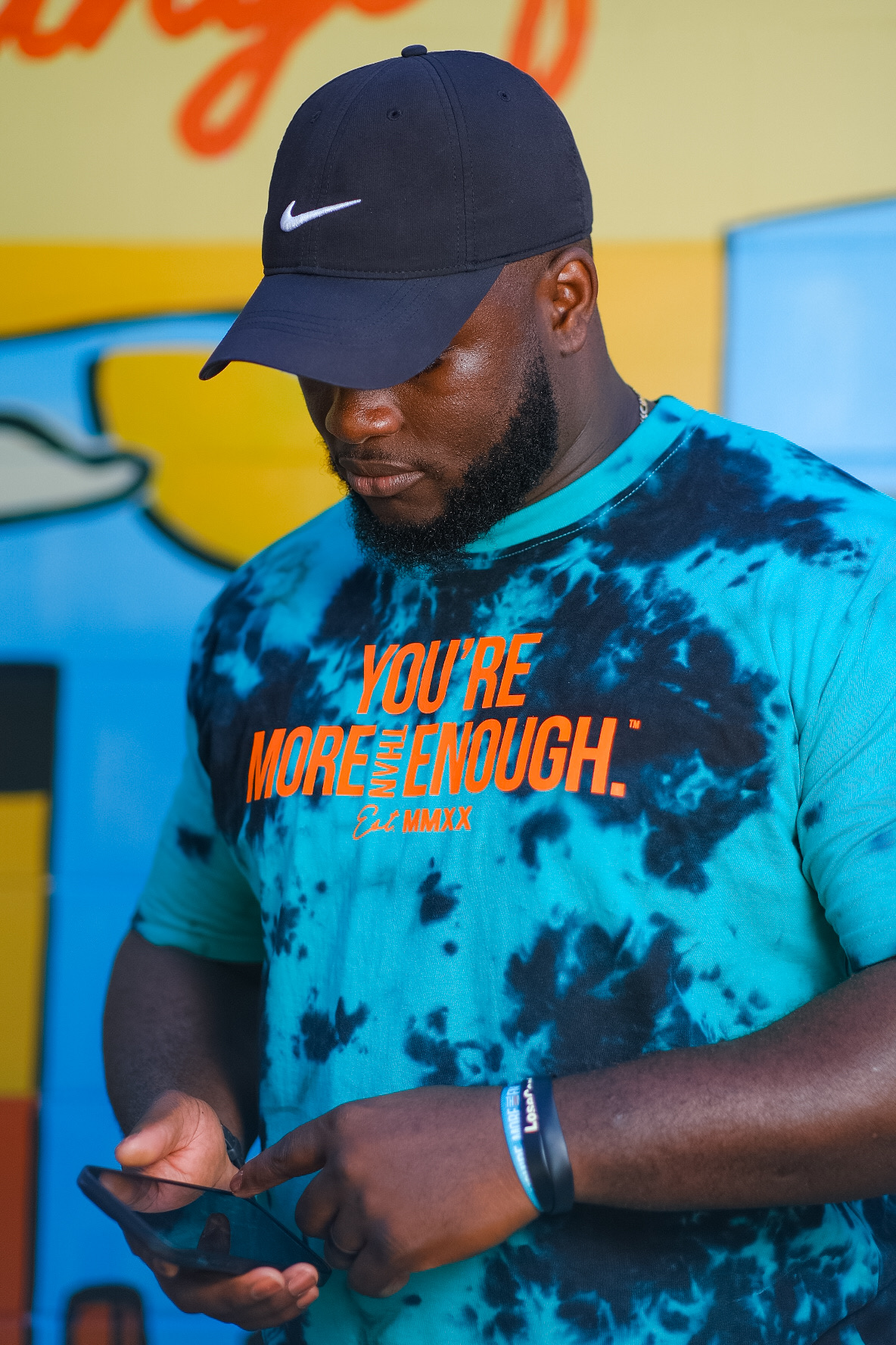 Man wearing a black Nike cap and blue tie-dye shirt looking at his phone, with a colorful background.