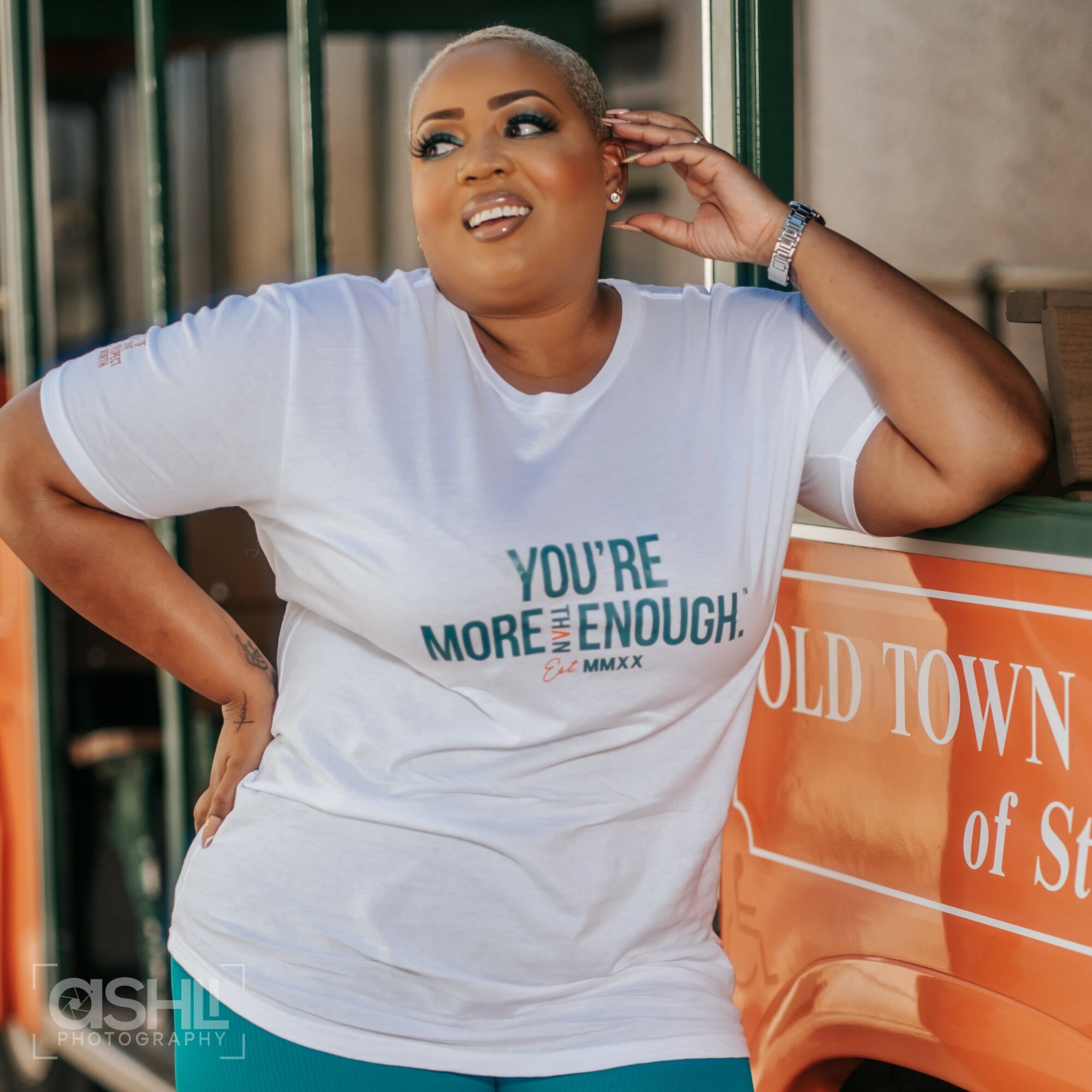 Woman with short hair and makeup, wearing a white T-shirt with text, standing outdoors near a green railing and orange sign.