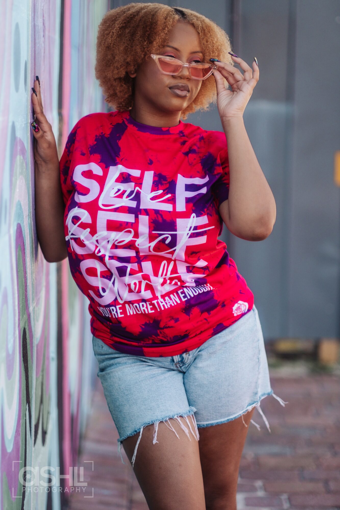 Young woman with curly hair wearing sunglasses, red and blue graphic T-shirt, and denim shorts, standing outdoors near a colorful wall.