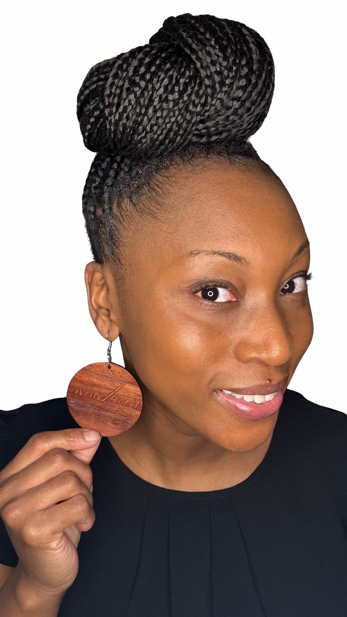 Woman with braided hair styled in a high bun, holding a large round earring, smiling, wearing a black top.