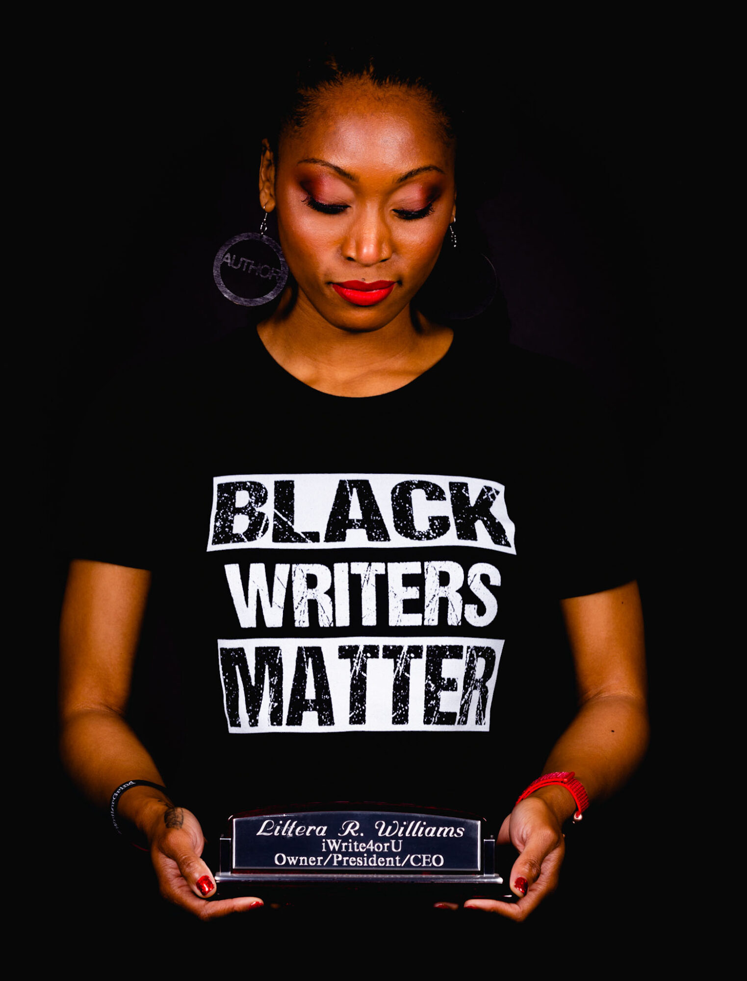 Woman with closed eyes holding a plaque, wearing a black T-shirt with white text, against a black background.