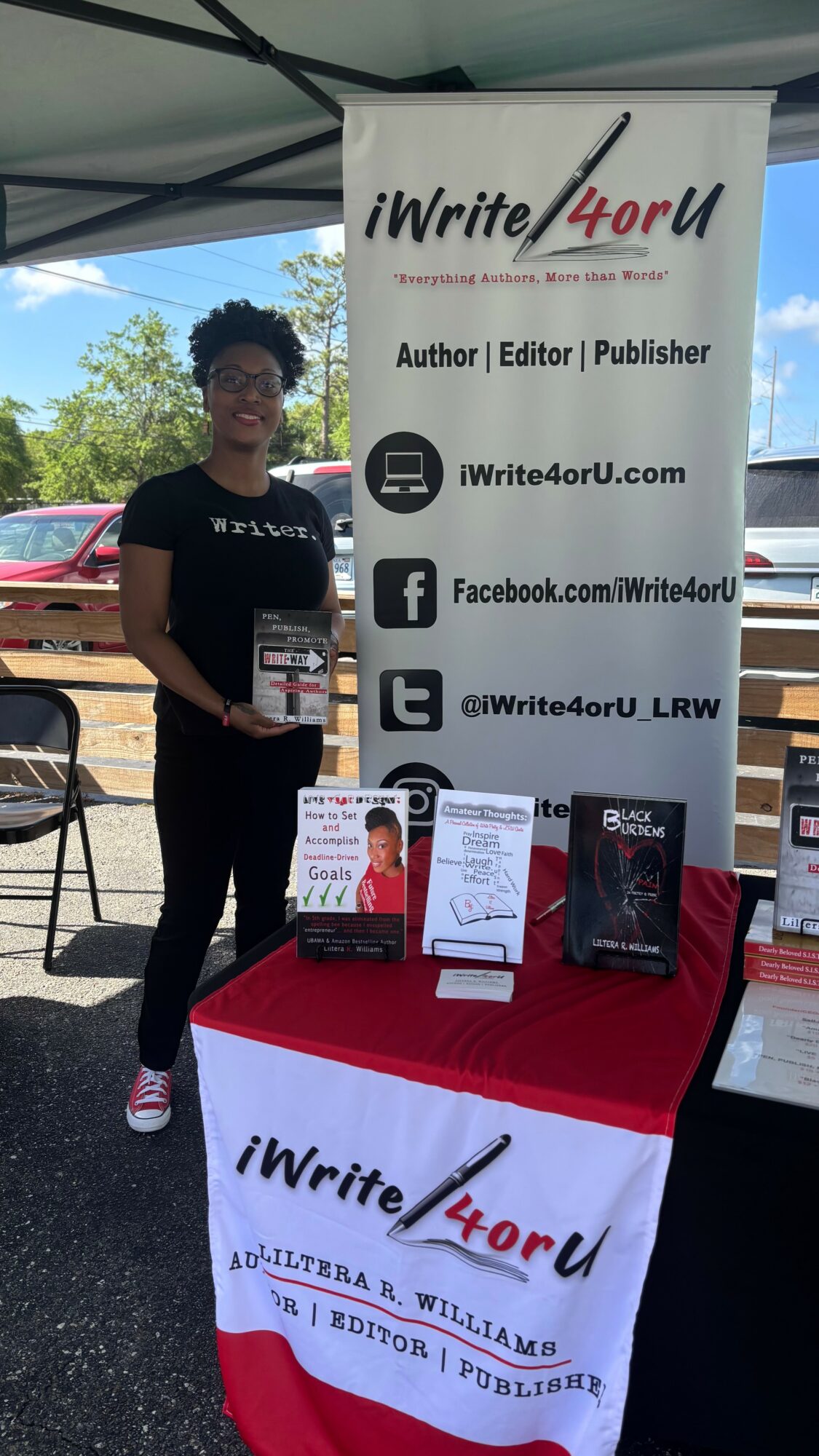 Person standing next to a table with books and a banner displaying 'iWrite4U' and social media info.