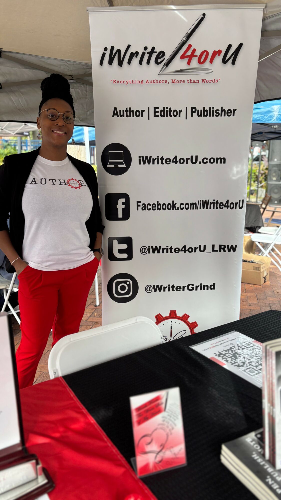 Woman standing next to a promotional banner at a booth, wearing a white t-shirt and red pants, smiling.