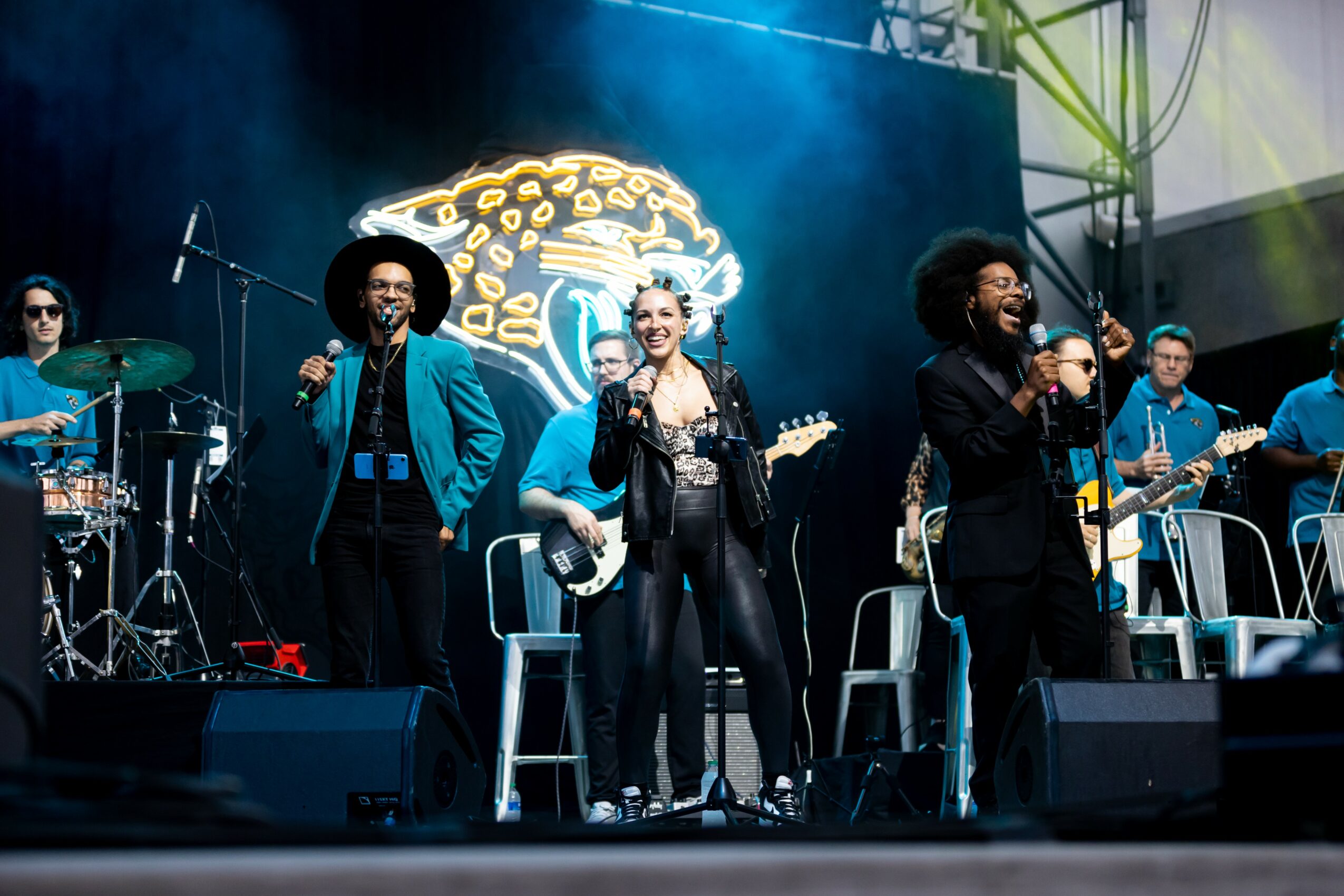 Group of musicians performing on stage with a large illuminated logo in the background, some singing and playing instruments.