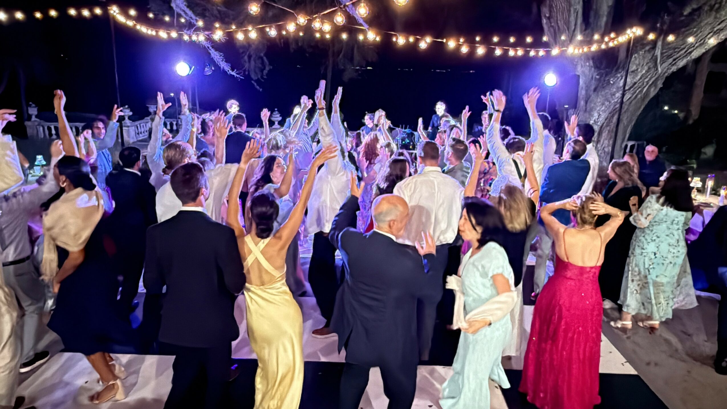 Crowd of people dancing outdoors at night under string lights, with some raising their hands, festive atmosphere.