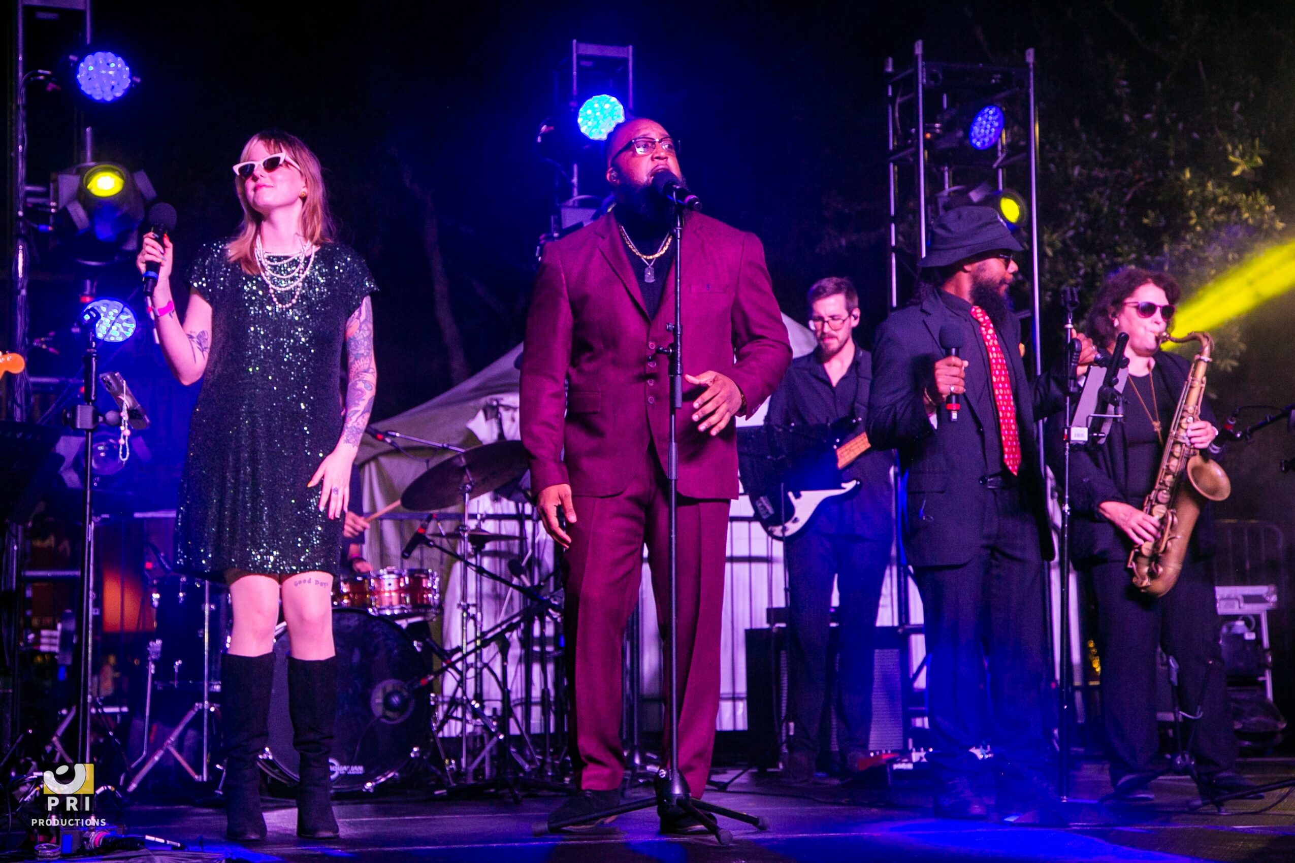 Band performing on stage with colorful lights, including a female singer, male singer, guitarist, saxophonist, and drummer.