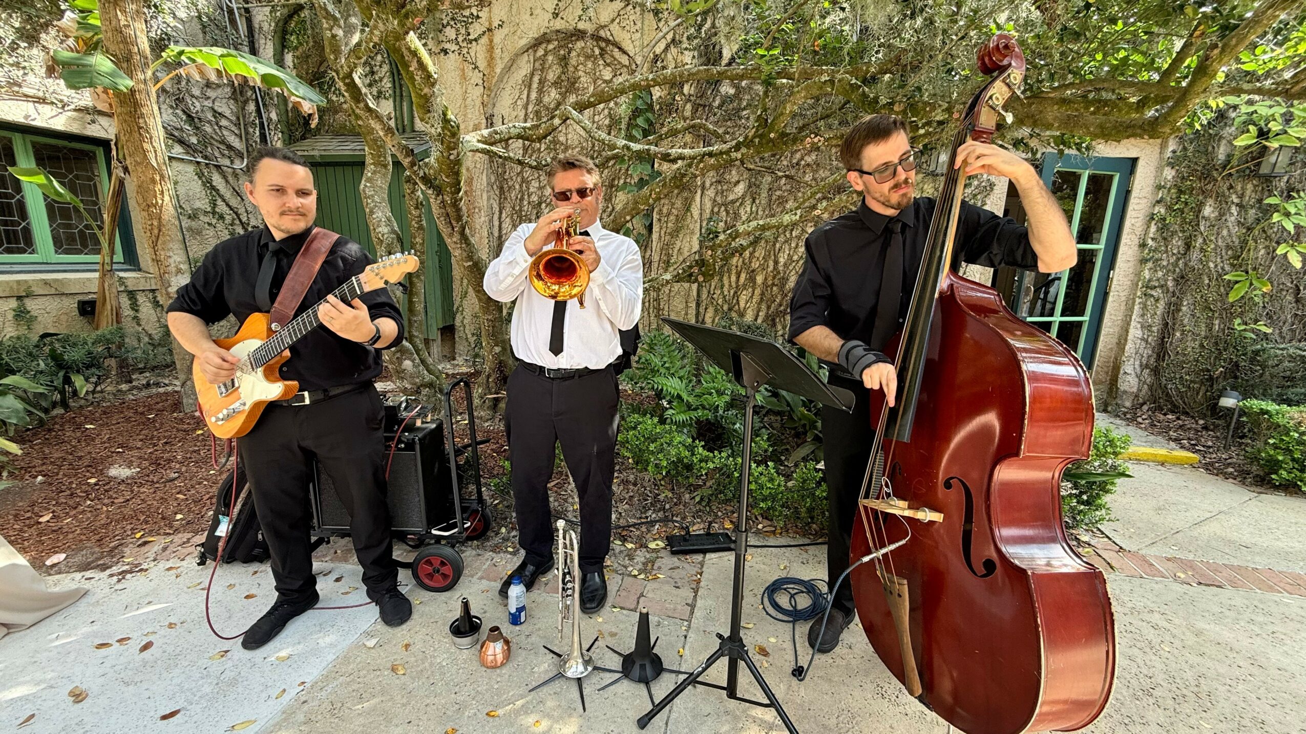Three musicians playing instruments outdoors, surrounded by trees and a building, with music stands and equipment nearby.
