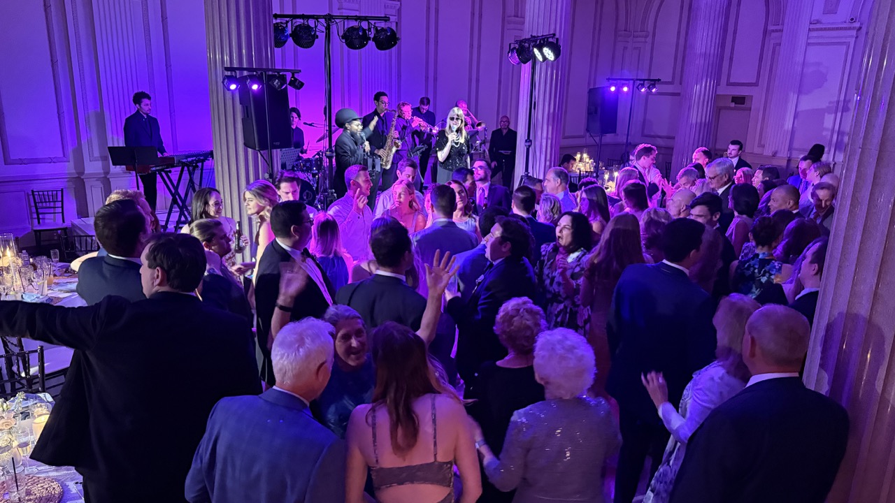 Crowd of people dancing and socializing at a formal event with purple lighting and a live band on stage.