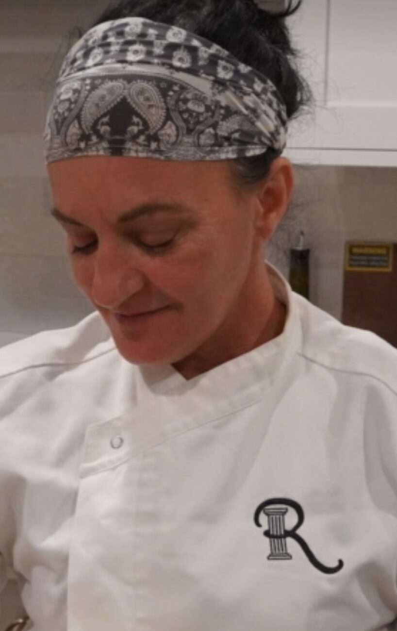 Woman wearing a patterned headscarf and white chef's coat, looking down, in a kitchen setting.