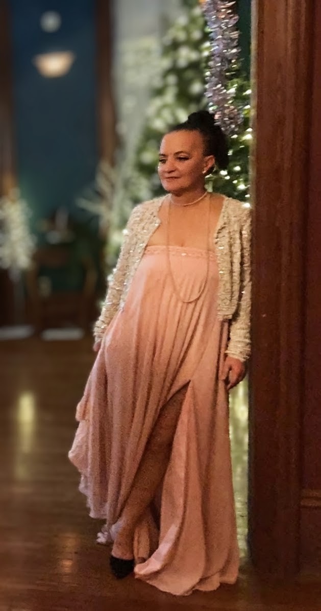 Woman in a peach dress and white jacket standing indoors near a decorated Christmas tree.