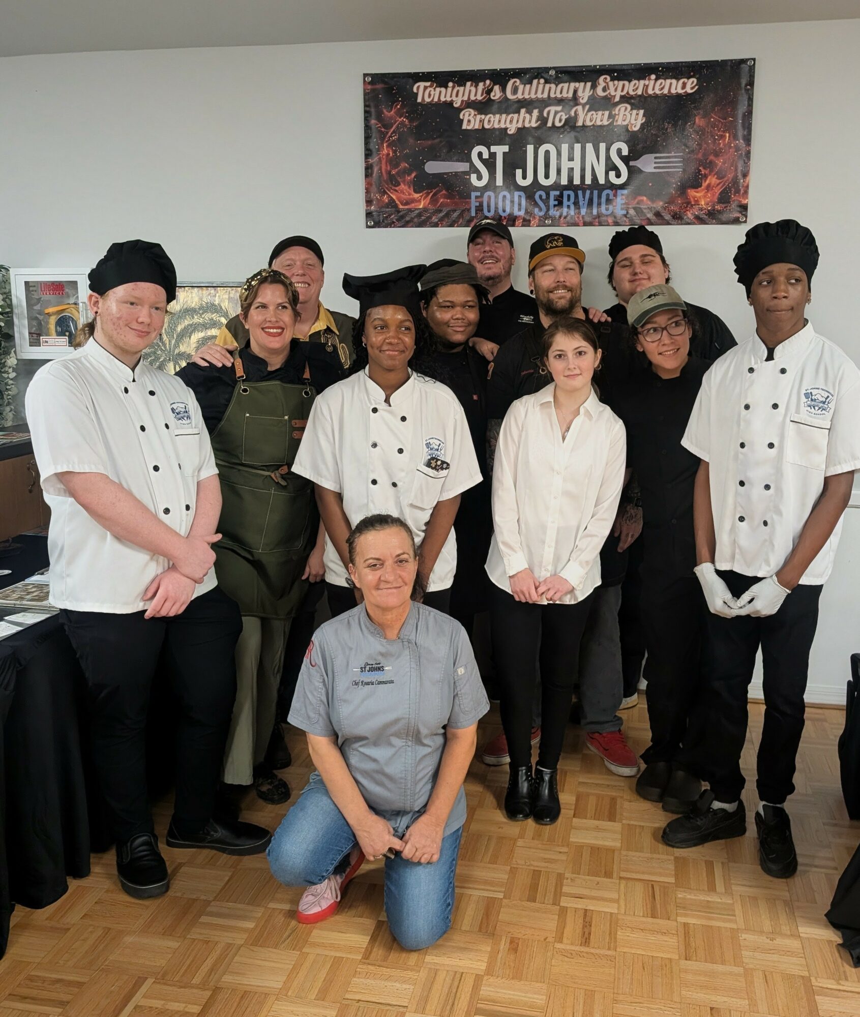 Group of people, including chefs and staff, standing indoors in front of a banner, some smiling, one kneeling in front.