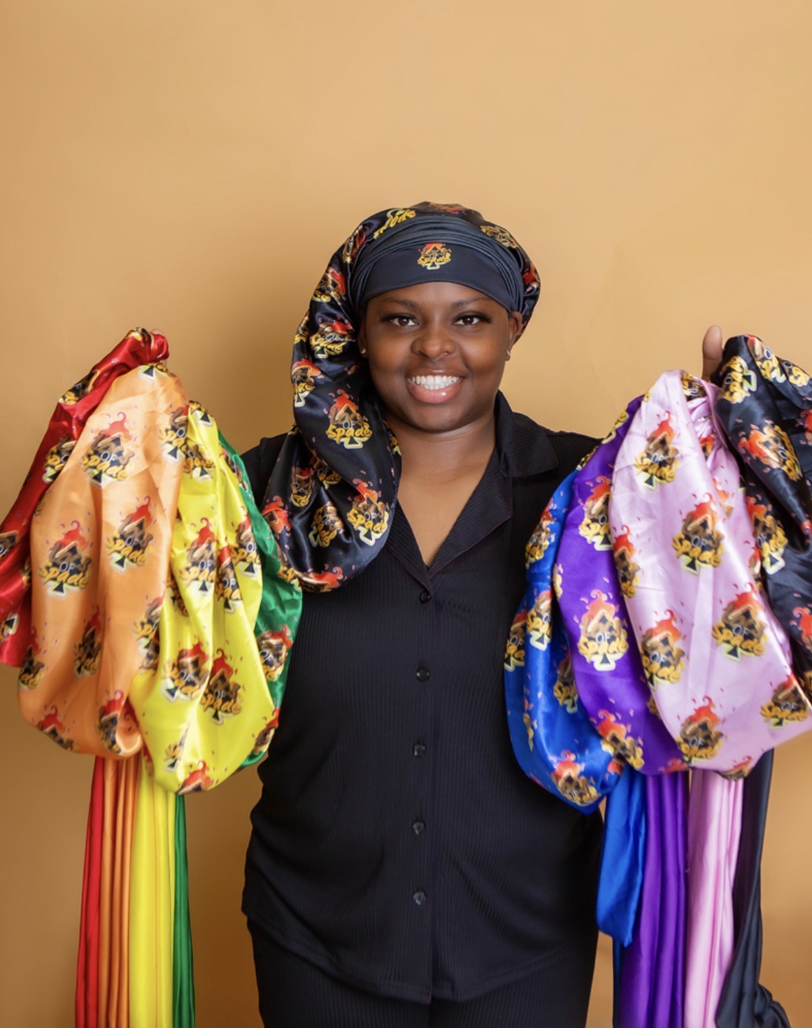 Smiling woman wearing a headscarf and black shirt holding colorful fabric bags against a beige background.