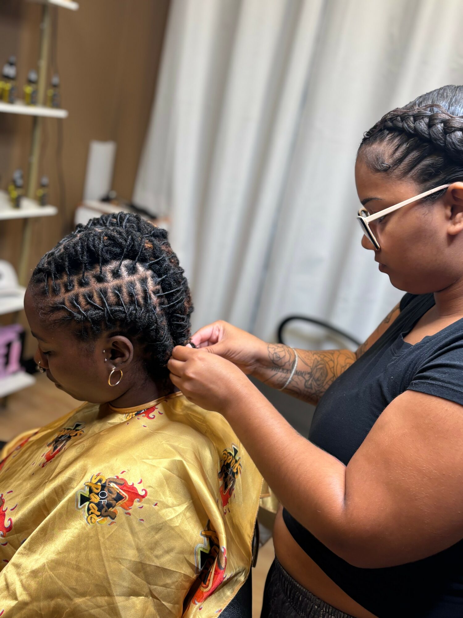 A hairstylist braiding a woman's hair in a salon, with shelves and curtains in the background.