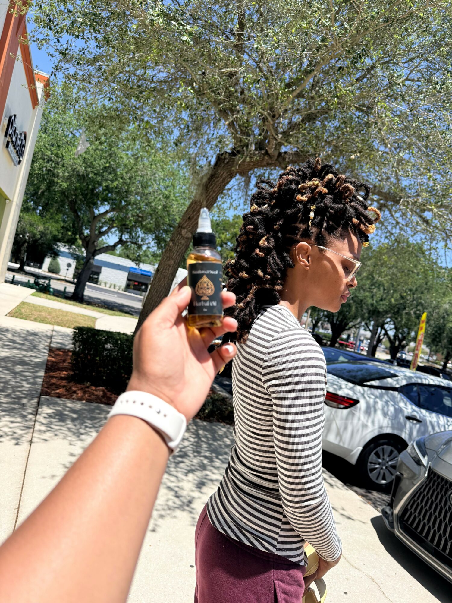 Person with curly hair and striped shirt outdoors, holding a bottle, trees and parked cars in background.