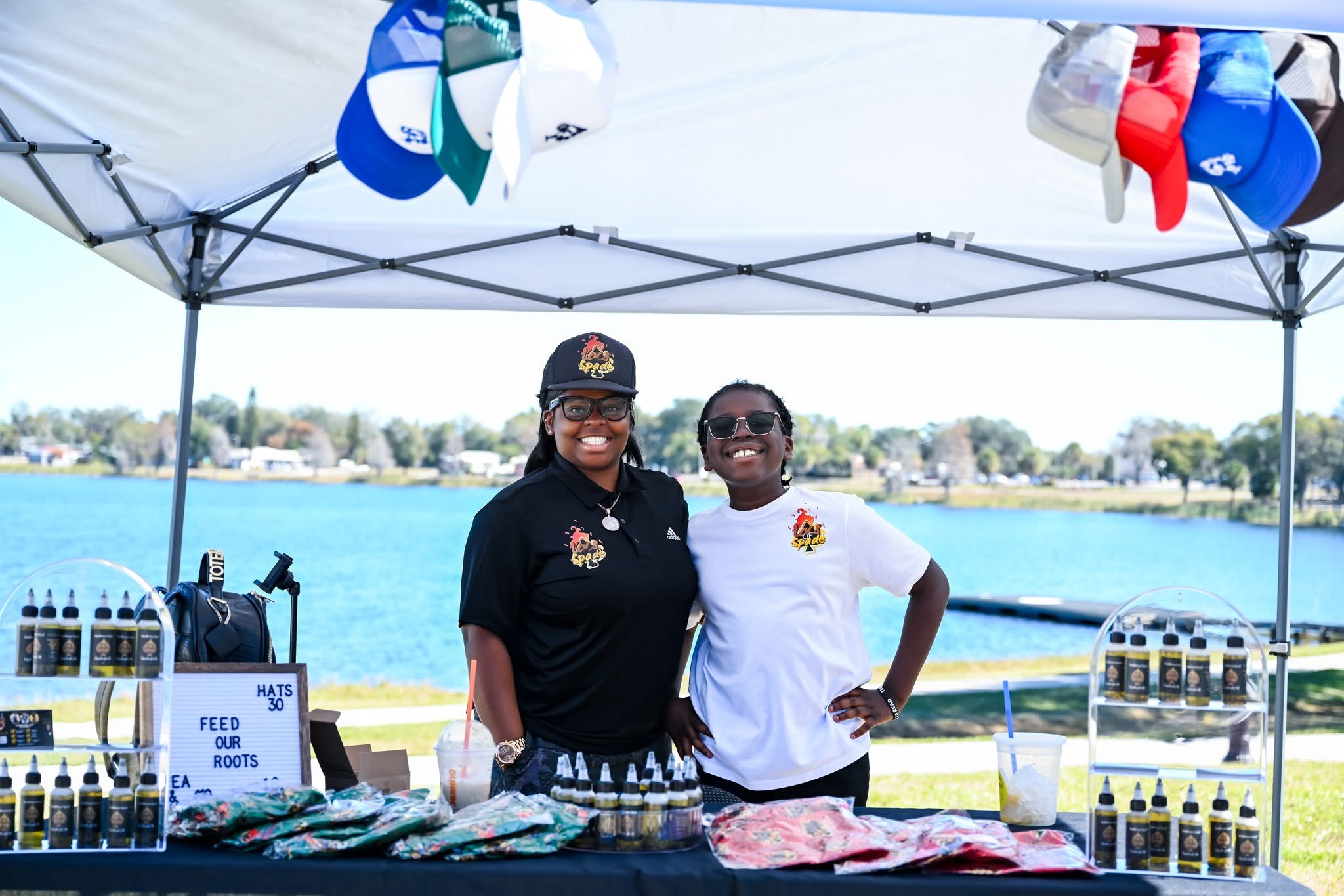 Two women stand under a canopy at a booth near a body of water, smiling, with bottles and items on the table.