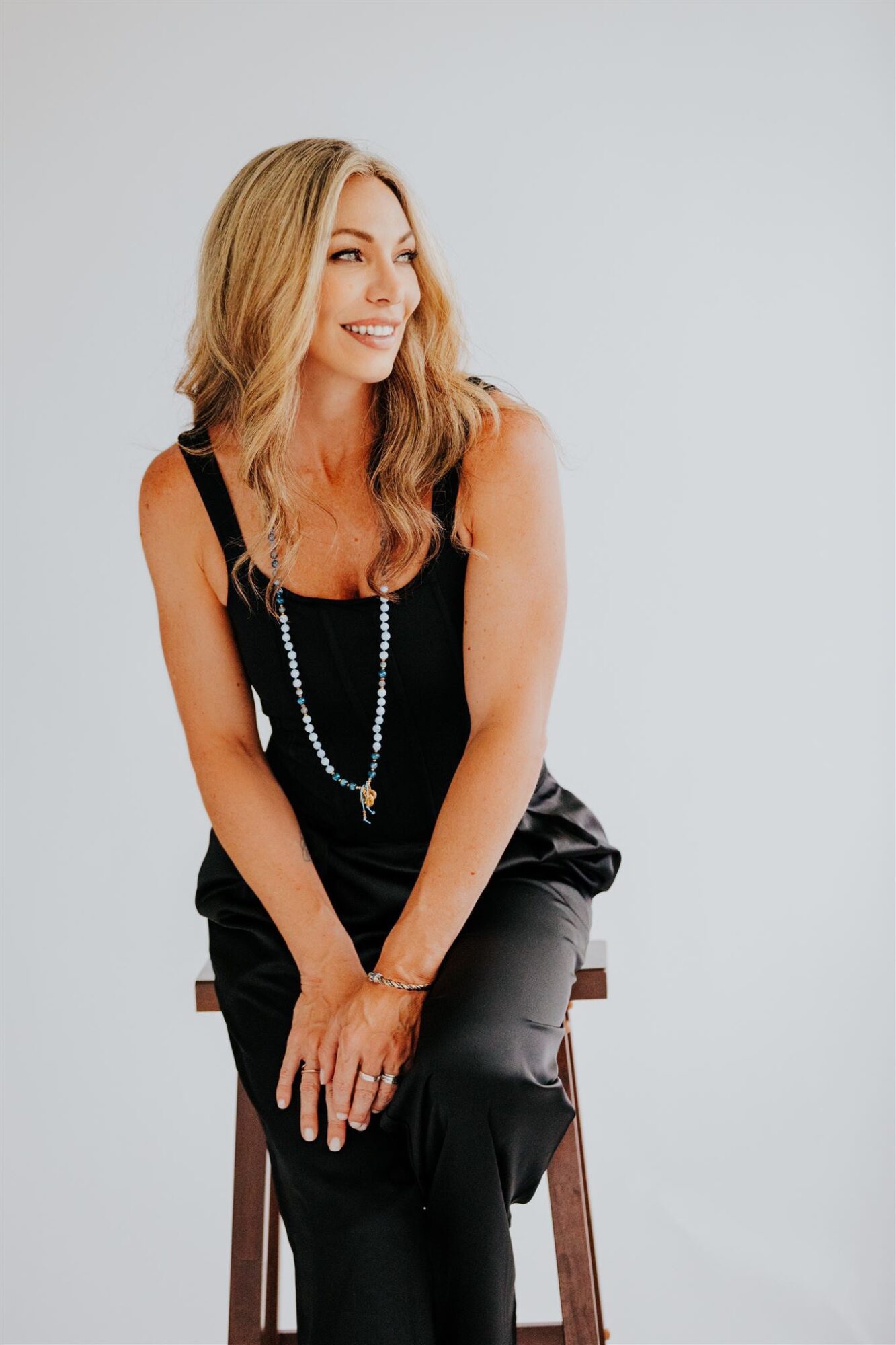 Woman with long blonde hair sitting on a stool, smiling, wearing a black dress and necklace, against a plain light background.