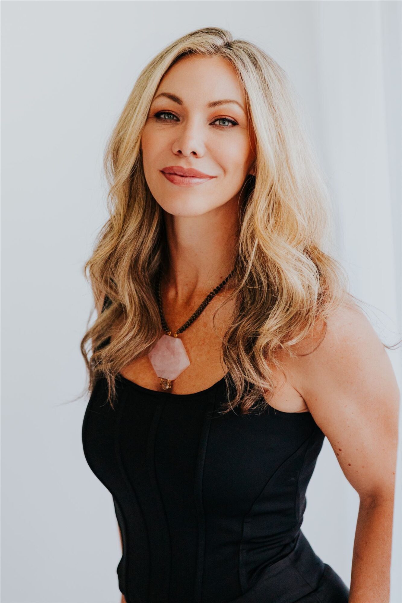 Woman with long blonde hair wearing a black top and pink necklace, smiling softly, standing indoors with white background.