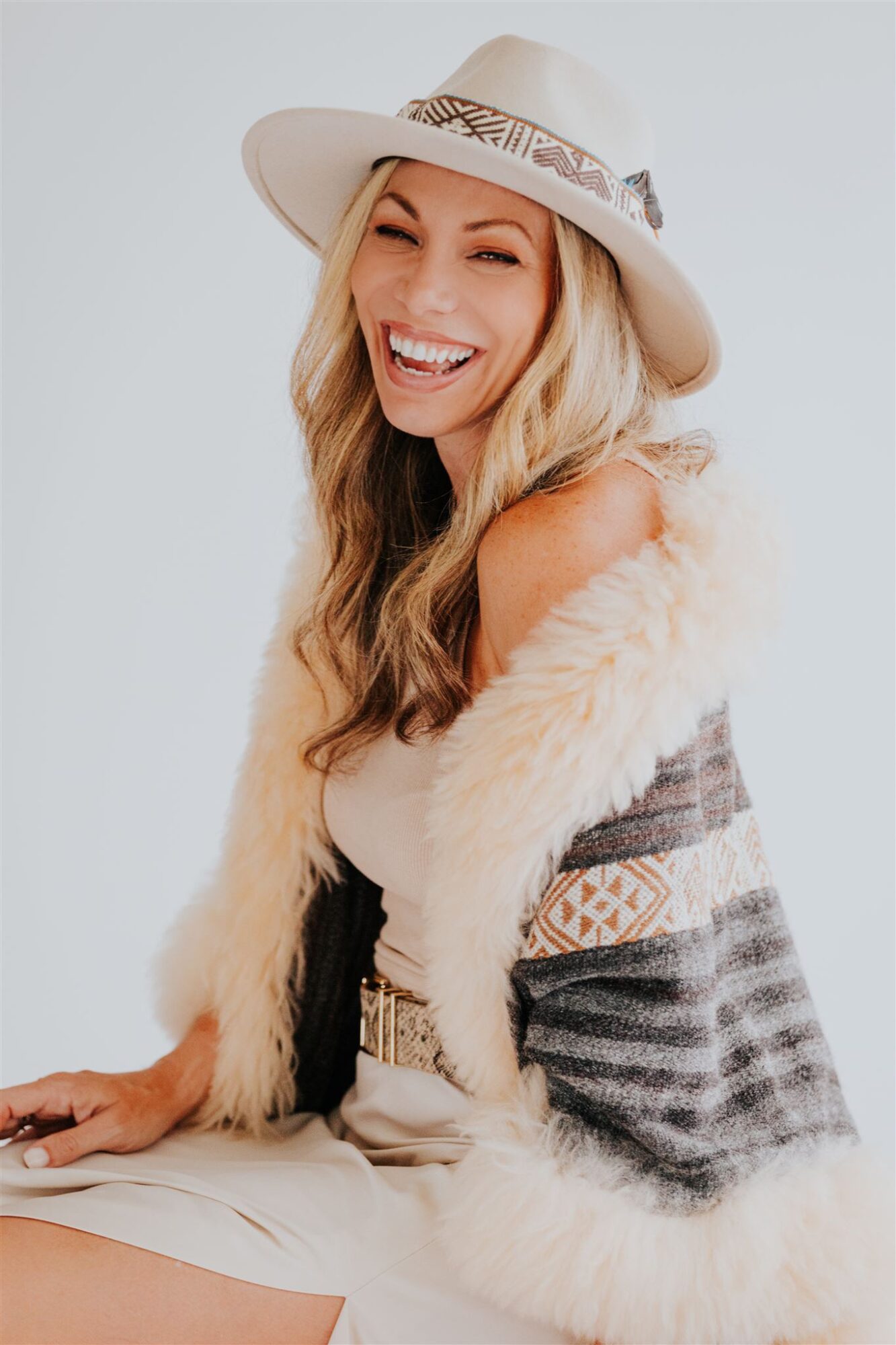Woman with long wavy hair wearing a wide-brimmed hat, smiling, dressed in a patterned shawl and light-colored dress.