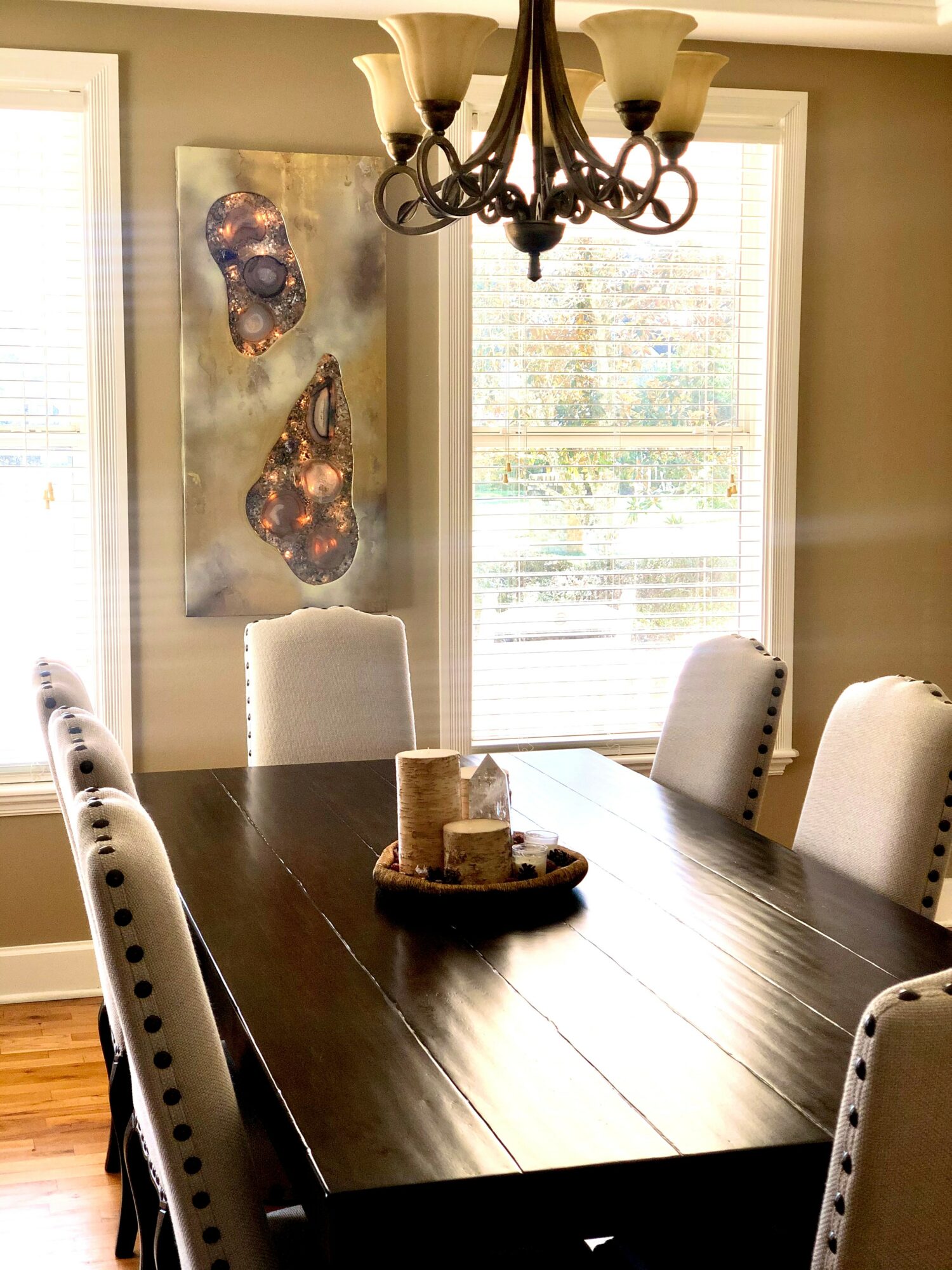 Dining room with a dark wooden table, six upholstered chairs, a chandelier, and a wall art piece, with sunlight through windows.