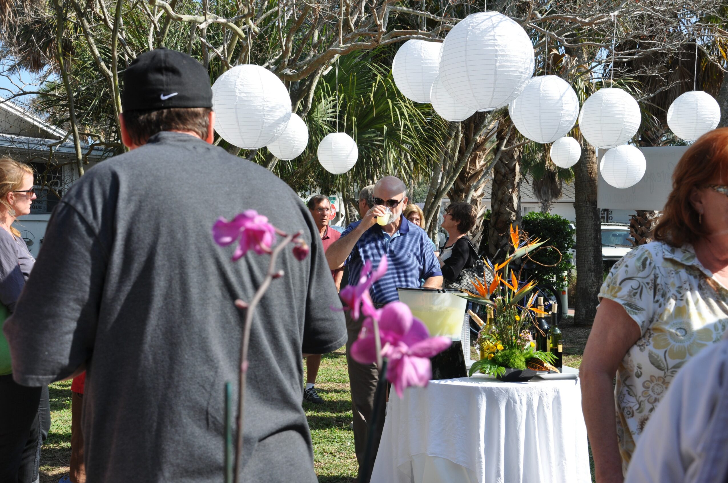 People gathered outdoors with white paper lanterns hanging above, some facing away, others talking near a table with flowers.