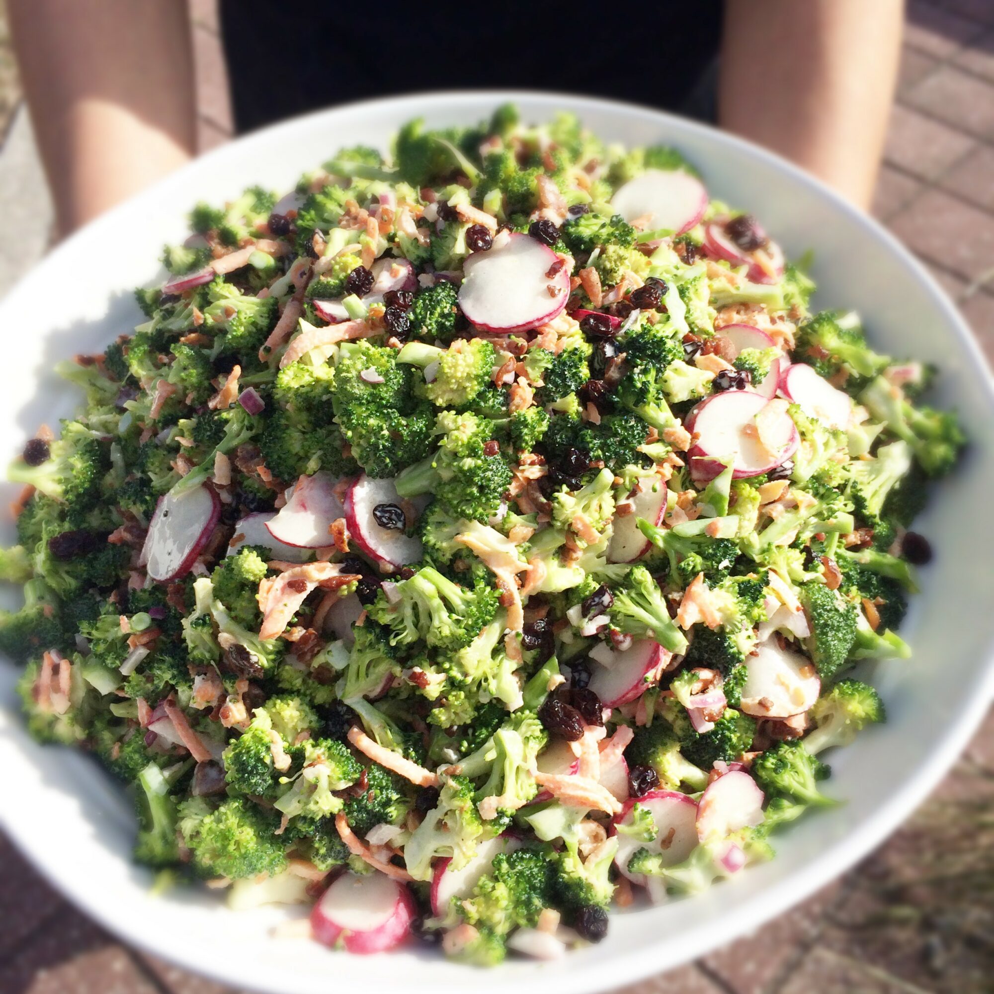 Large bowl of broccoli, radish slices, and other vegetables, held outdoors with sunlight, in a white dish.