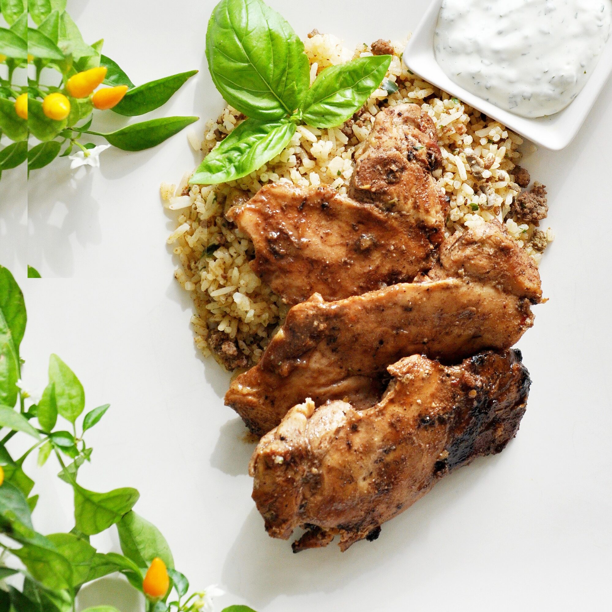 Cooked chicken pieces served with rice, garnished with basil leaves, and a side of white sauce, on a white plate.