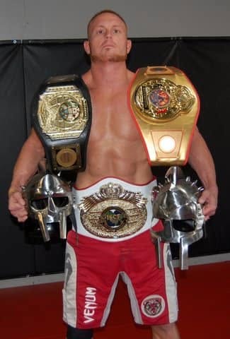 Man with two championship belts over shoulders and a third belt around waist, standing shirtless in a gym.
