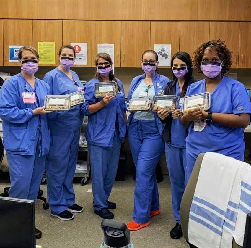 Group of six healthcare workers in blue scrubs and purple masks holding trays in a hospital room.