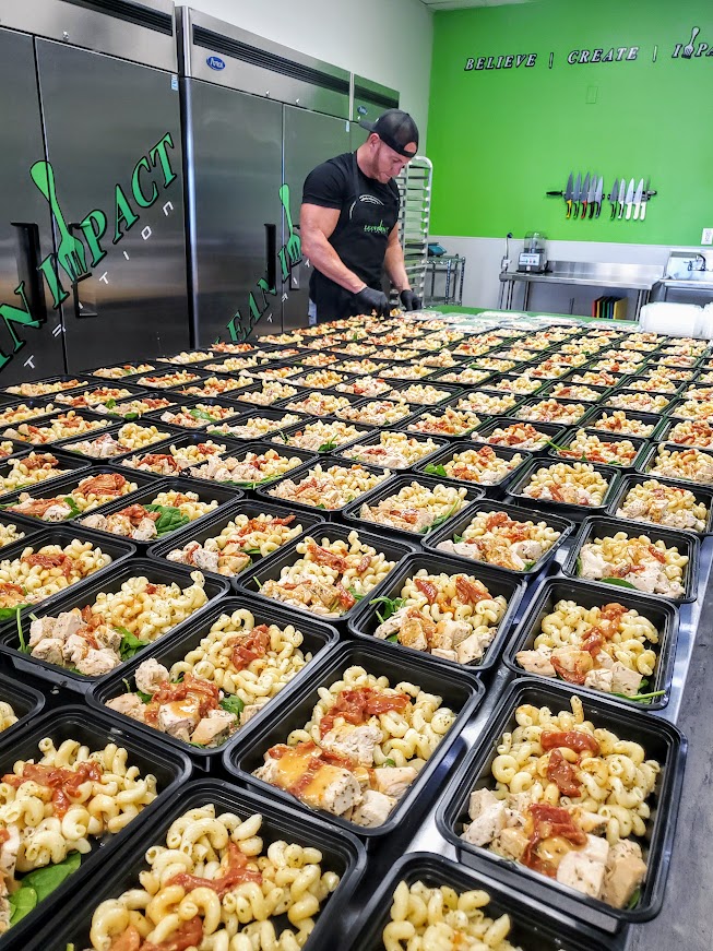 Person preparing multiple black containers filled with pasta and toppings in a bright green kitchen.