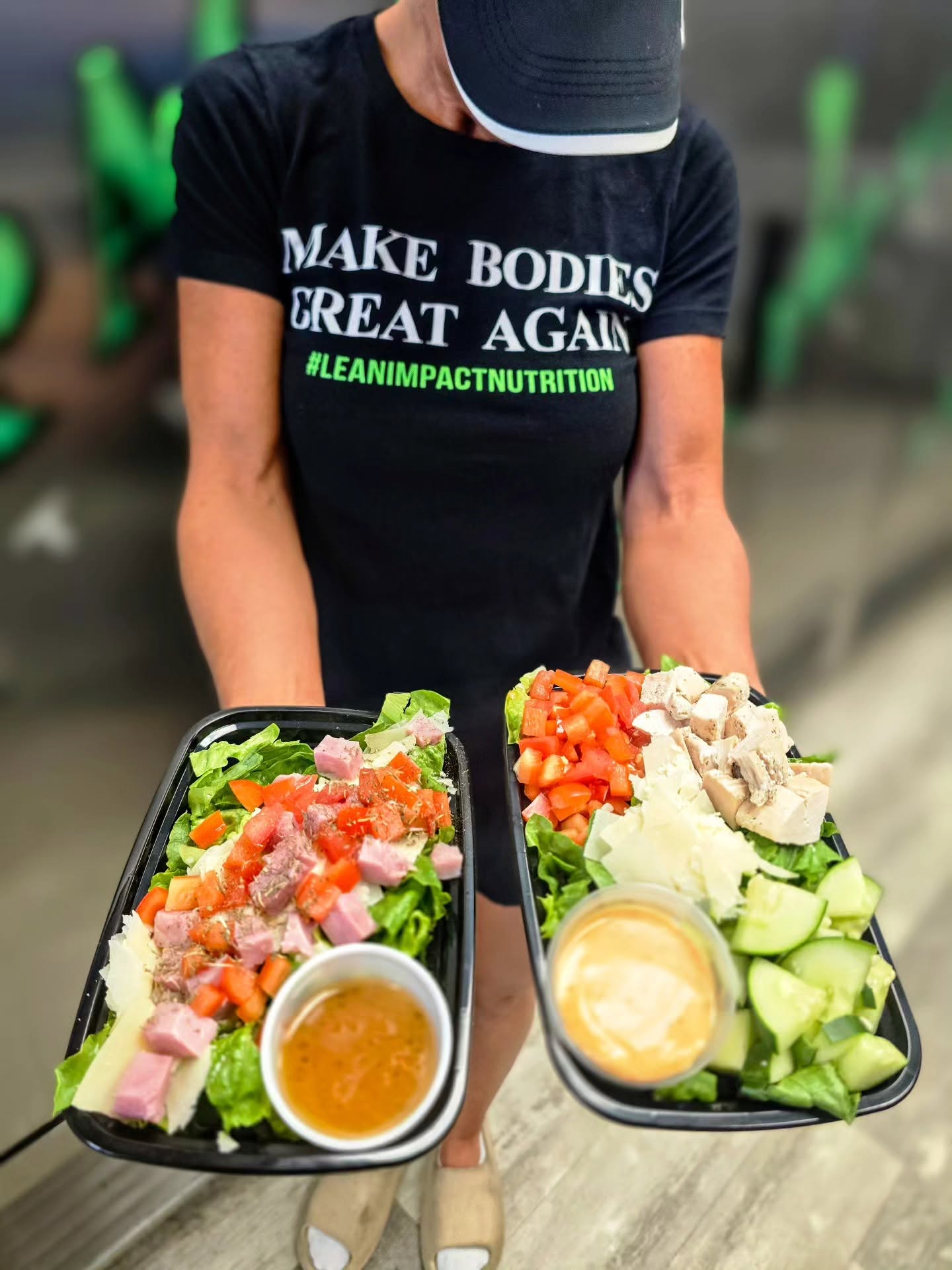 Person wearing a black shirt and cap holding two trays of salad with vegetables and dressing, blurred background.