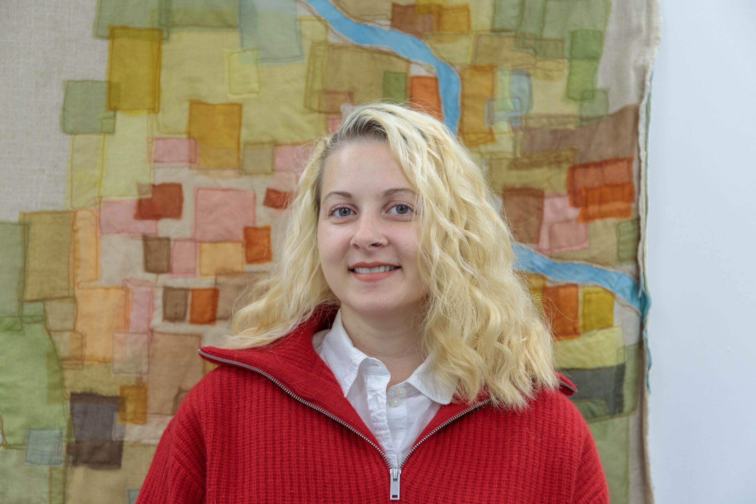 Young woman with blonde curly hair smiling, wearing a red jacket and white shirt, standing in front of a colorful abstract background.