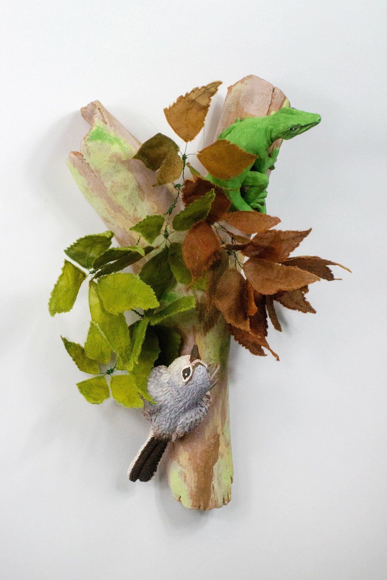A small bird figurine perched on a branch with green and brown leaves, against a white background.