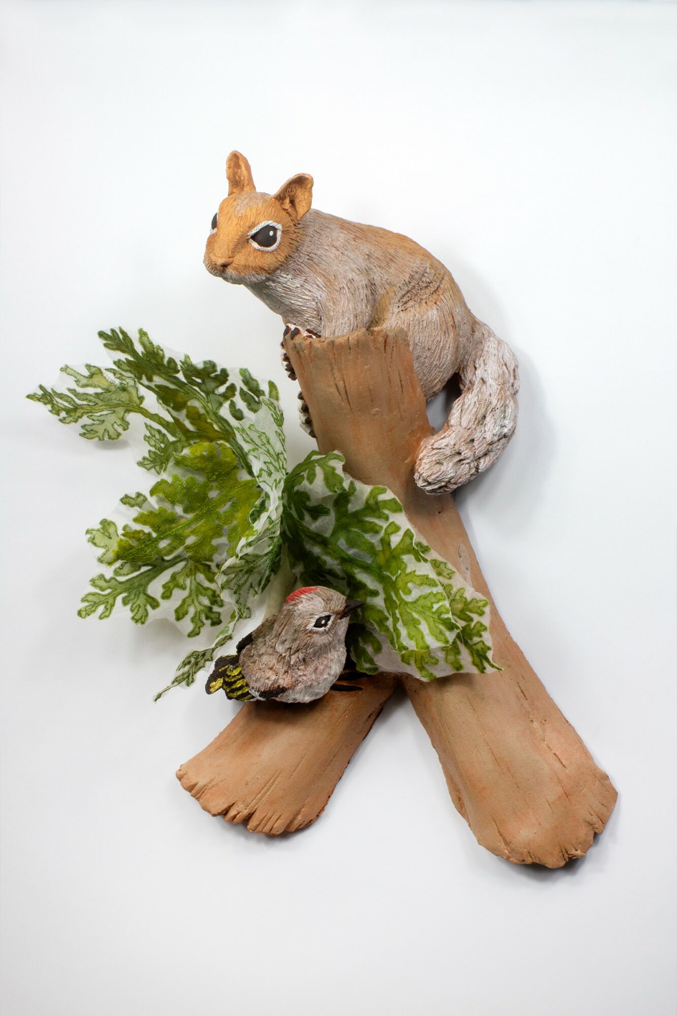 A squirrel figurine perched on a wooden branch with green leaves, against a white background.