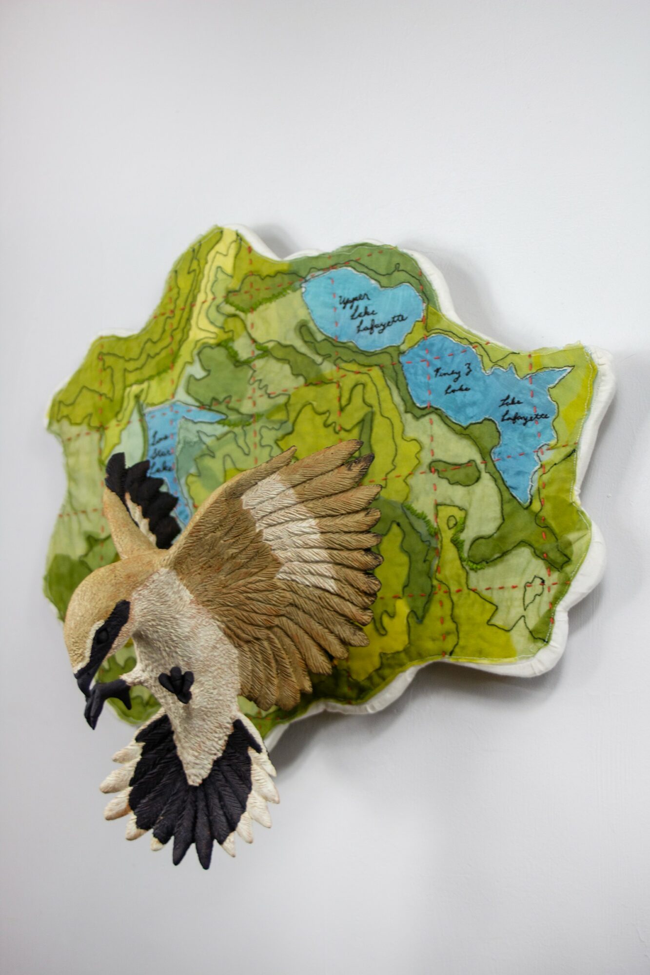 Fabric map with a bird embroidery, showing lakes and land features, on a white background.
