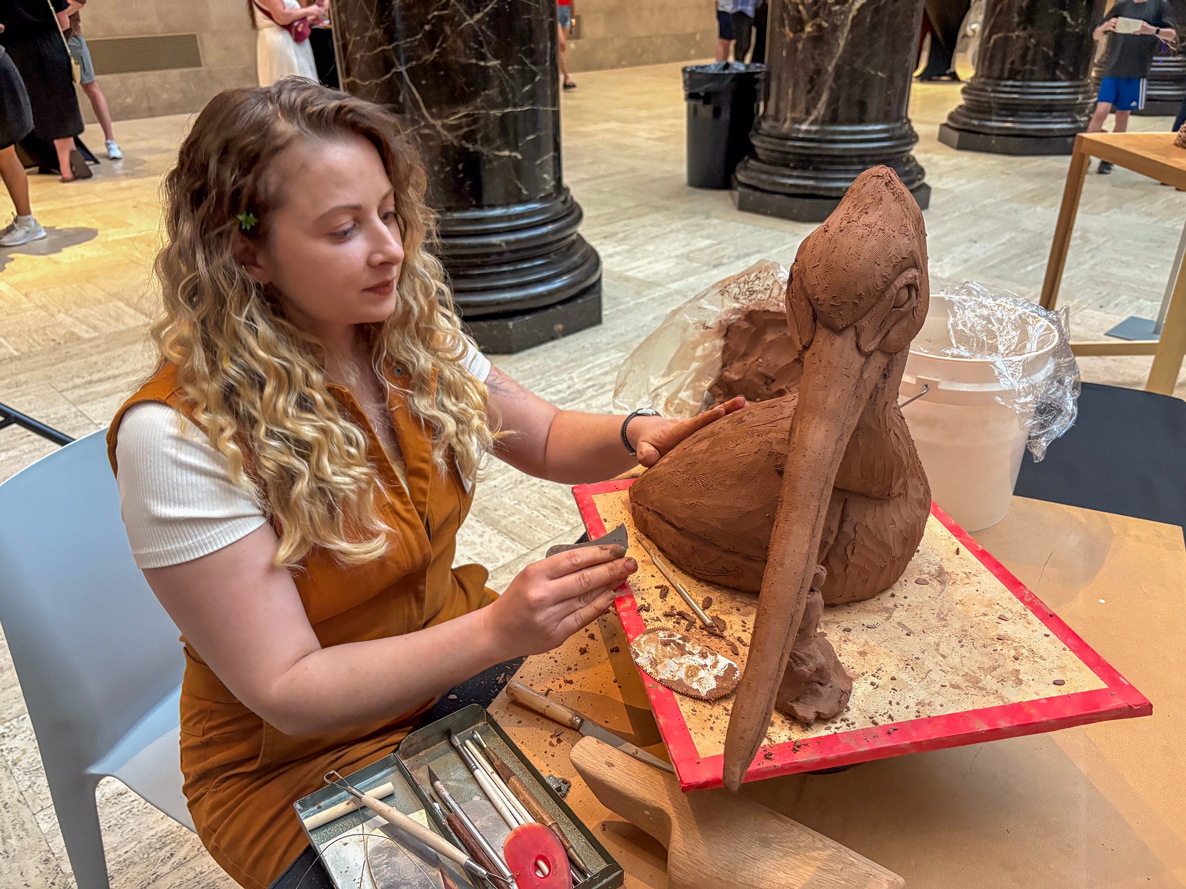 Young woman with curly hair sitting at a table, working on a large wooden sculpture in a public space.
