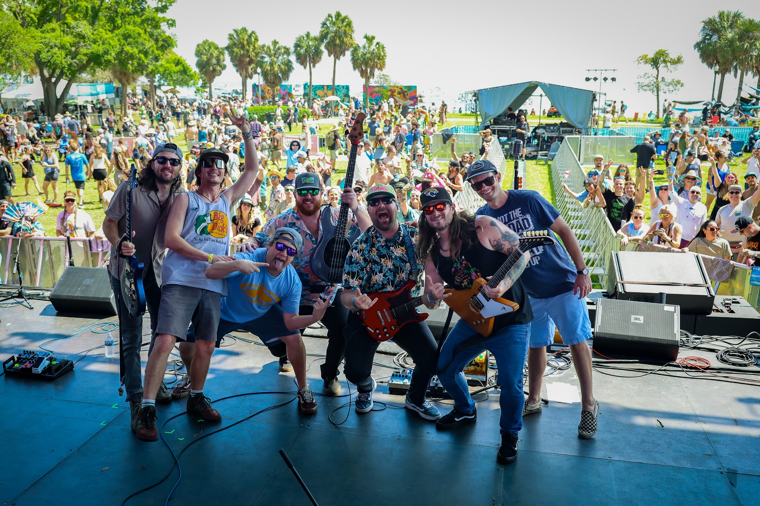 Group of six musicians performing on stage at outdoor event with large crowd and trees in background.