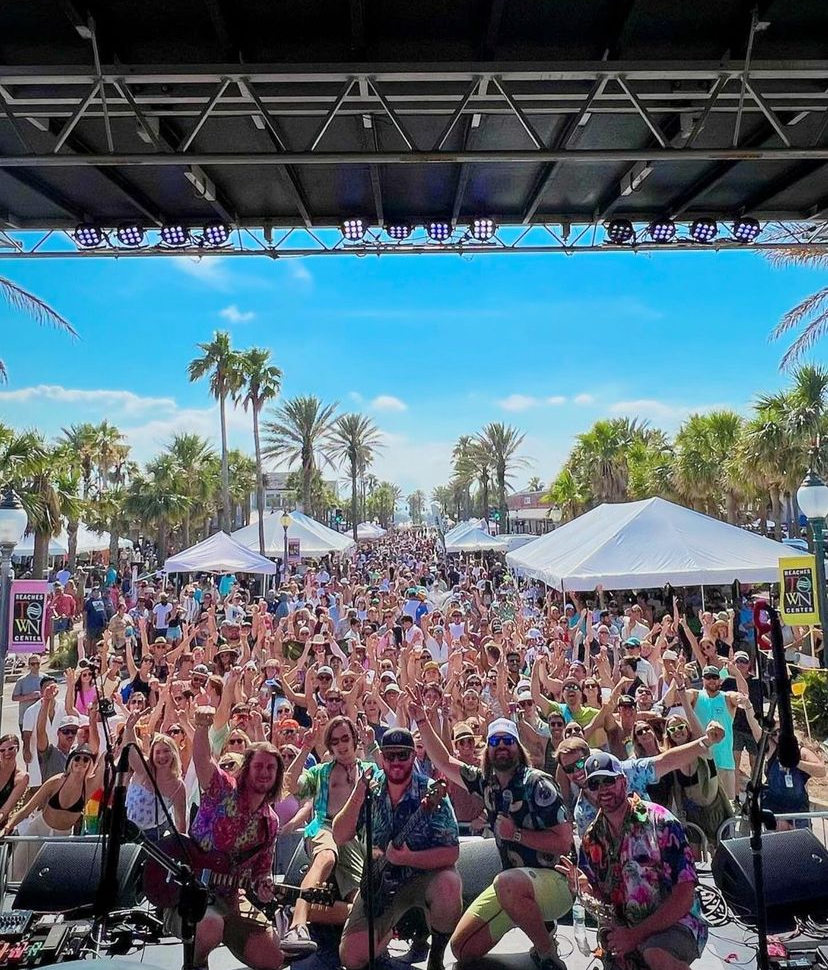 Crowd of people outdoors with palm trees, tents, and a blue sky, raising hands at an event, viewed from stage.