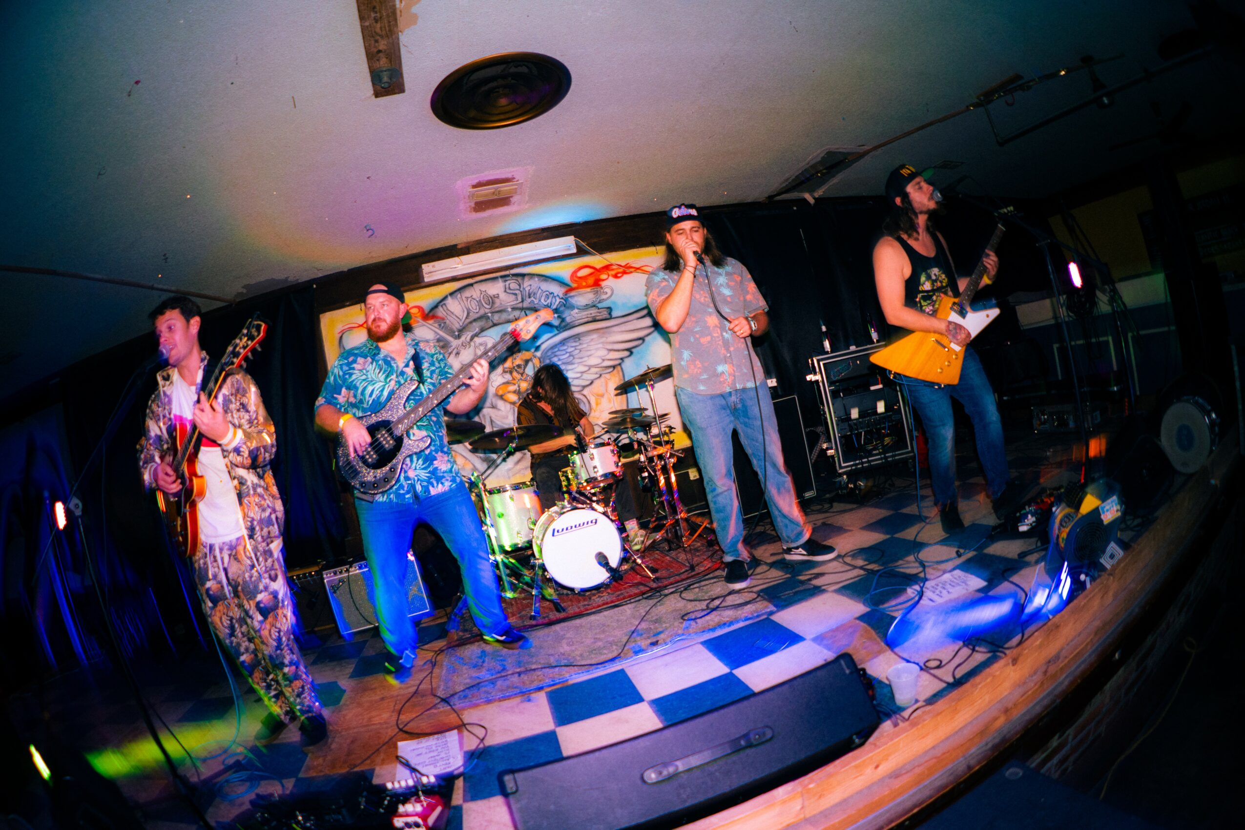 Band performing on stage with five members, colorful backdrop, and checkered floor, playing instruments and singing.