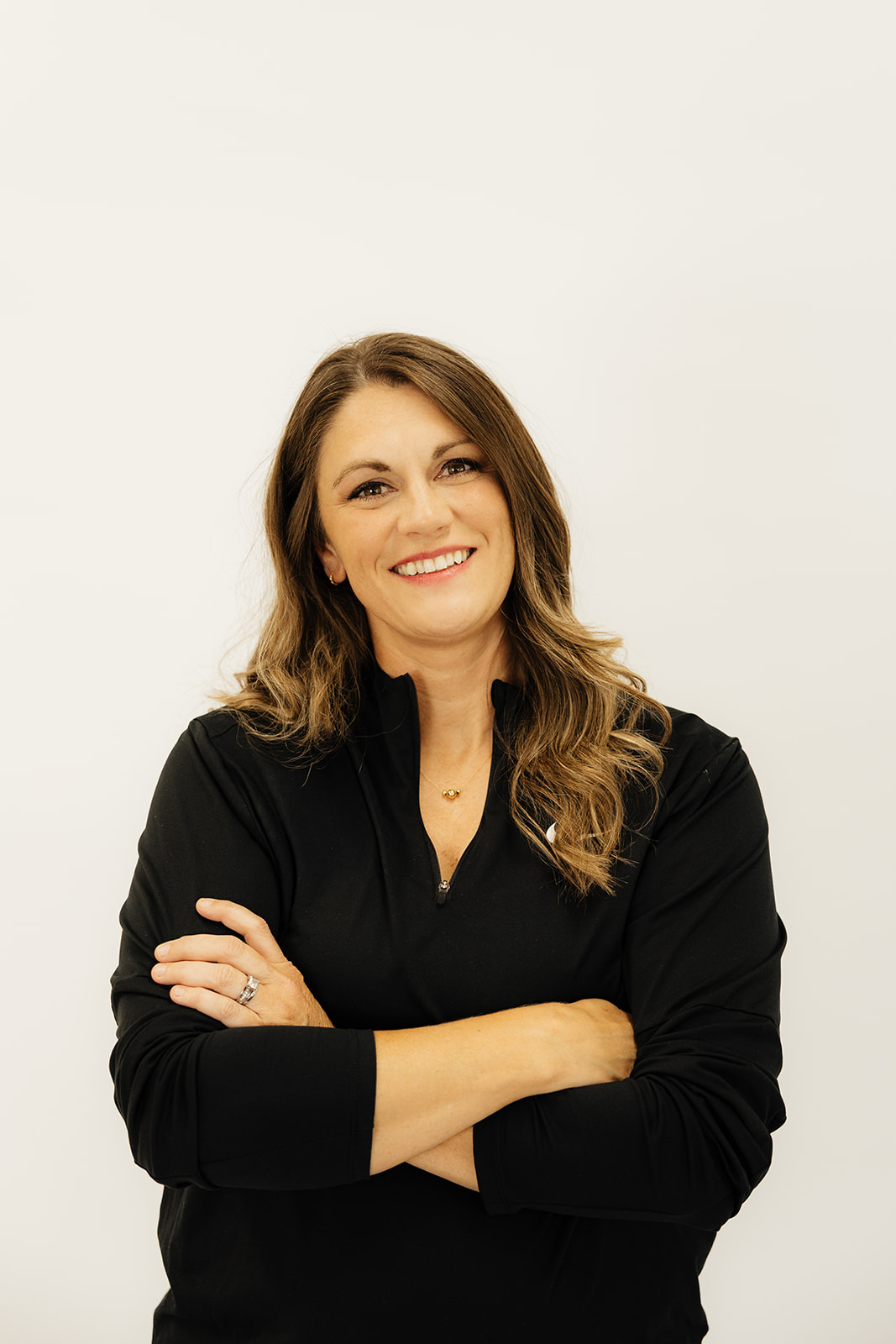 Woman with long wavy hair smiling, arms crossed, wearing a black top, standing against a plain white background.